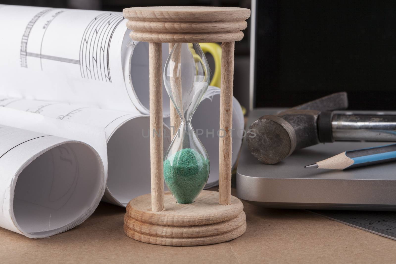 Architectural plans rolled with silver laptop hamer and a pencil and green hourglass