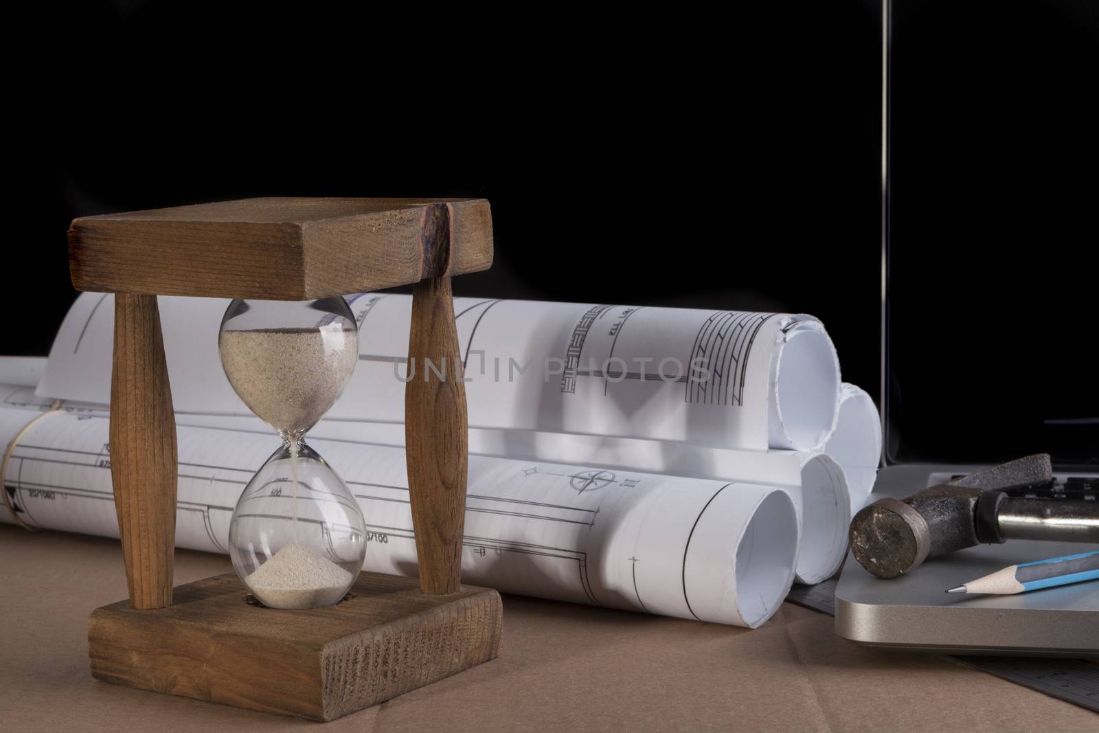 Architectural plans rolled with silver laptop hammer and a pencil and old wood hourglass