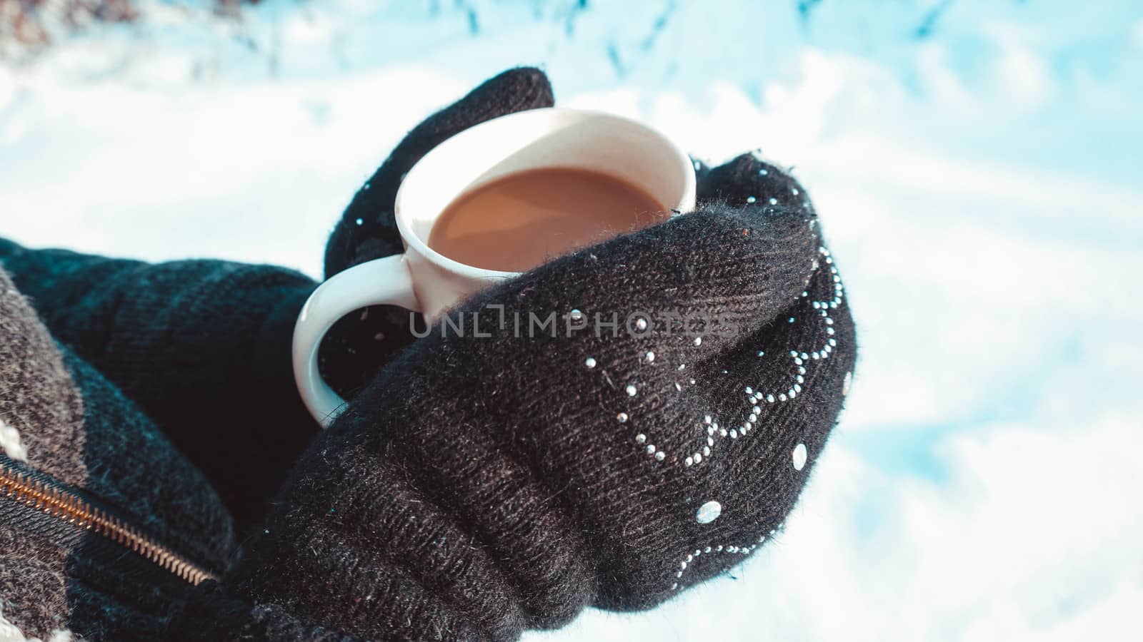 Warm cup of hot coffee warming in the hands in mittens. Womens hands on the background of snow, winter