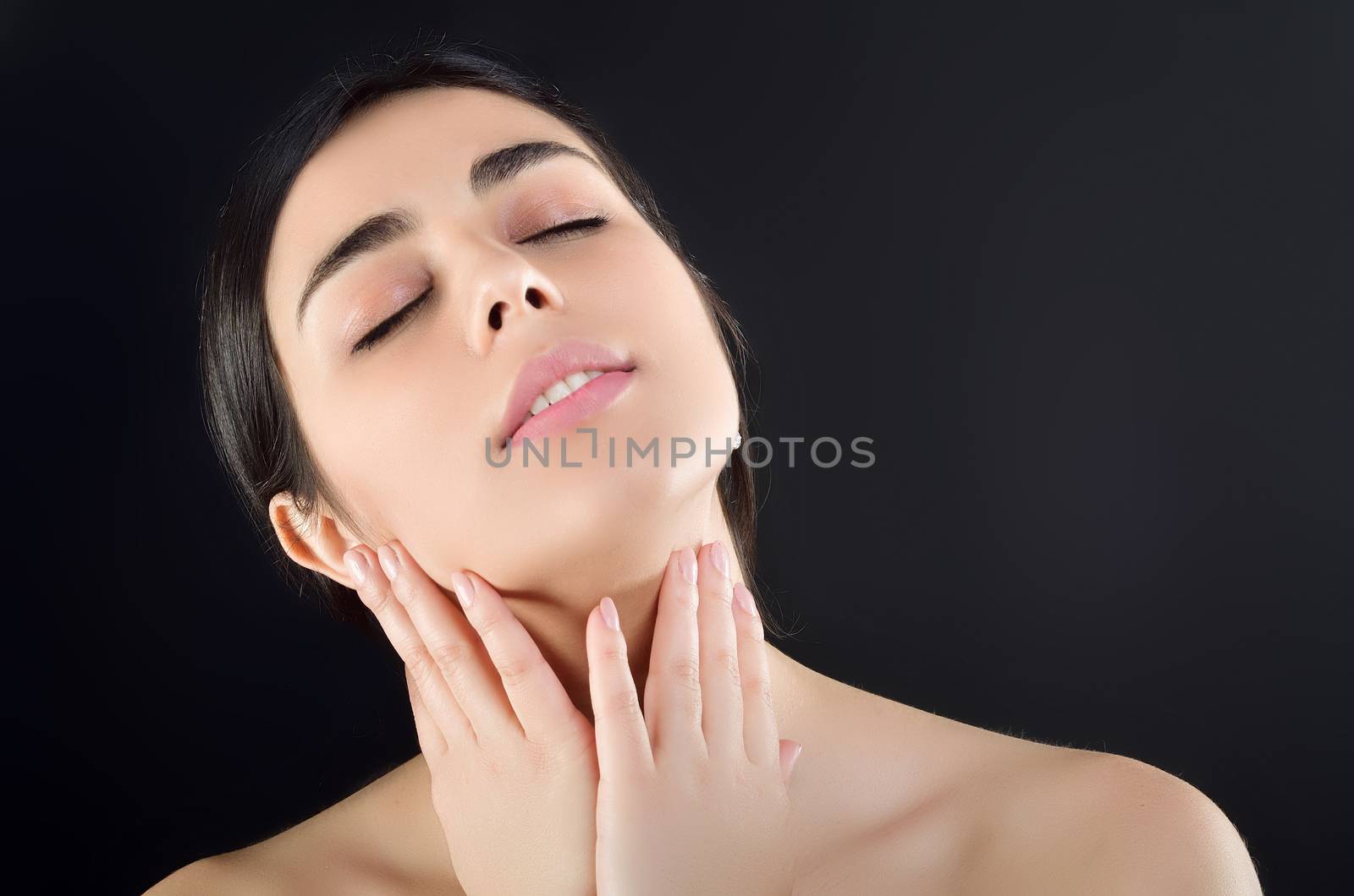 Pretty woman gently runs her fingers around her neck with her eyes closed, turning her head up. Studio photography, black background