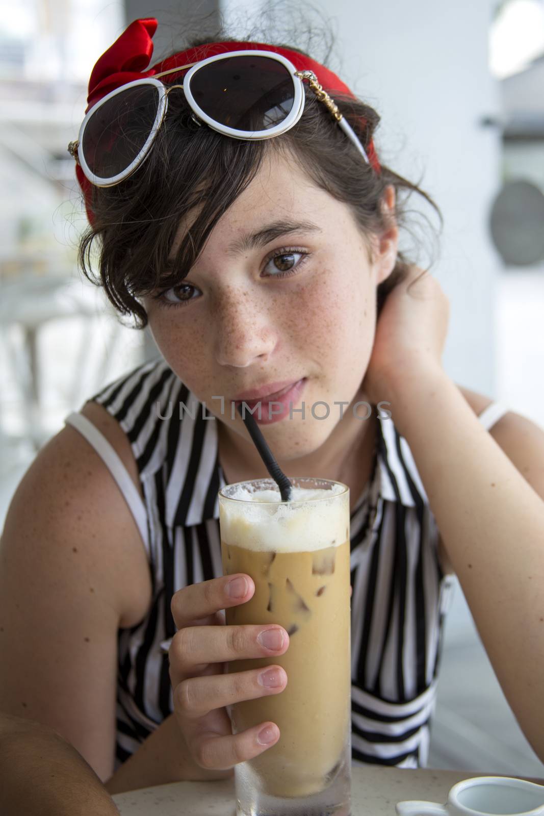 A teenage girl with pretty freckles drinks ice coffee in a cafe near the window. by Anelik