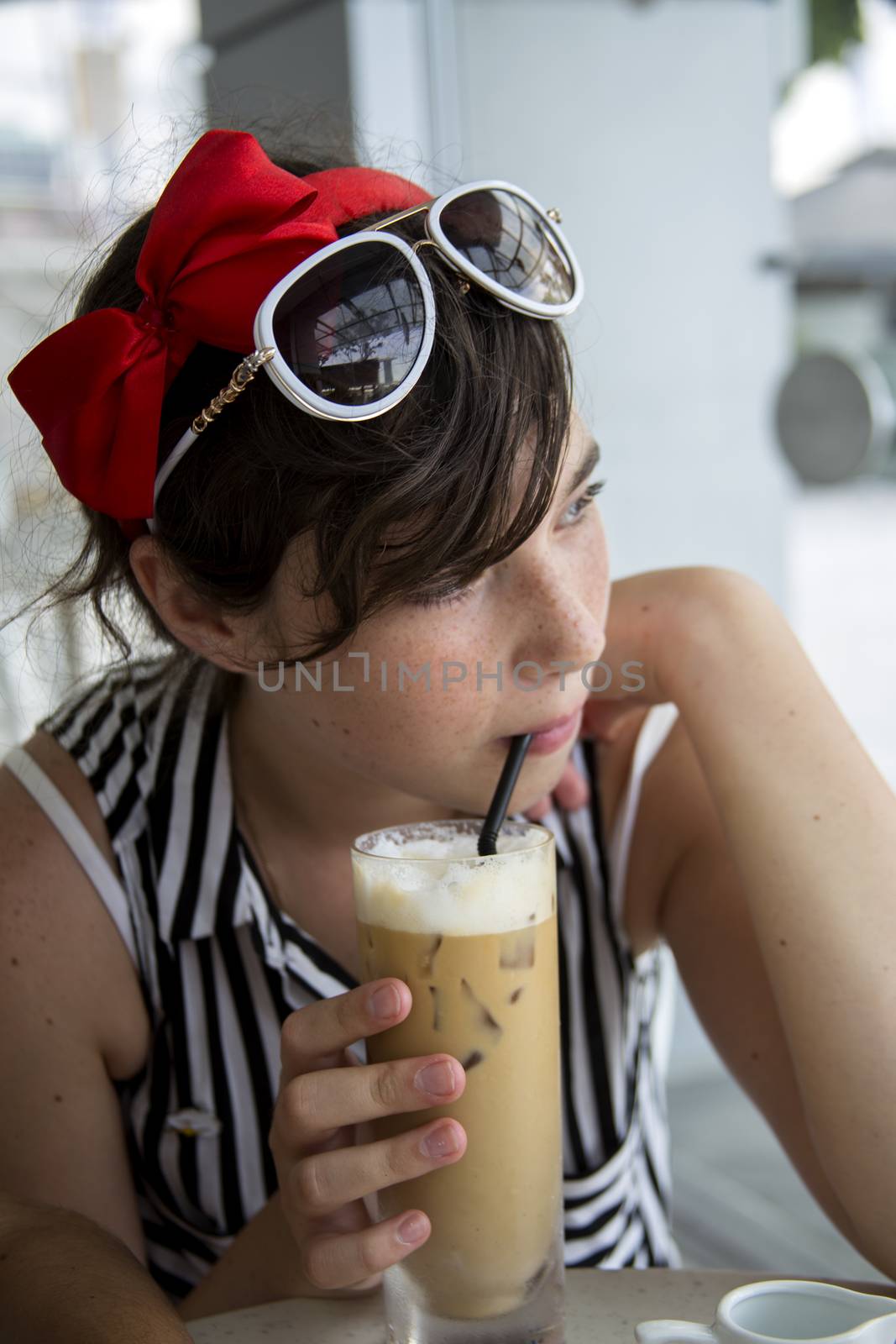 A teenage girl with pretty freckles on her face, dressed in a summer blouse and with sun glasses on her head, drinks ice coffee through a straw in a cafe near the window.