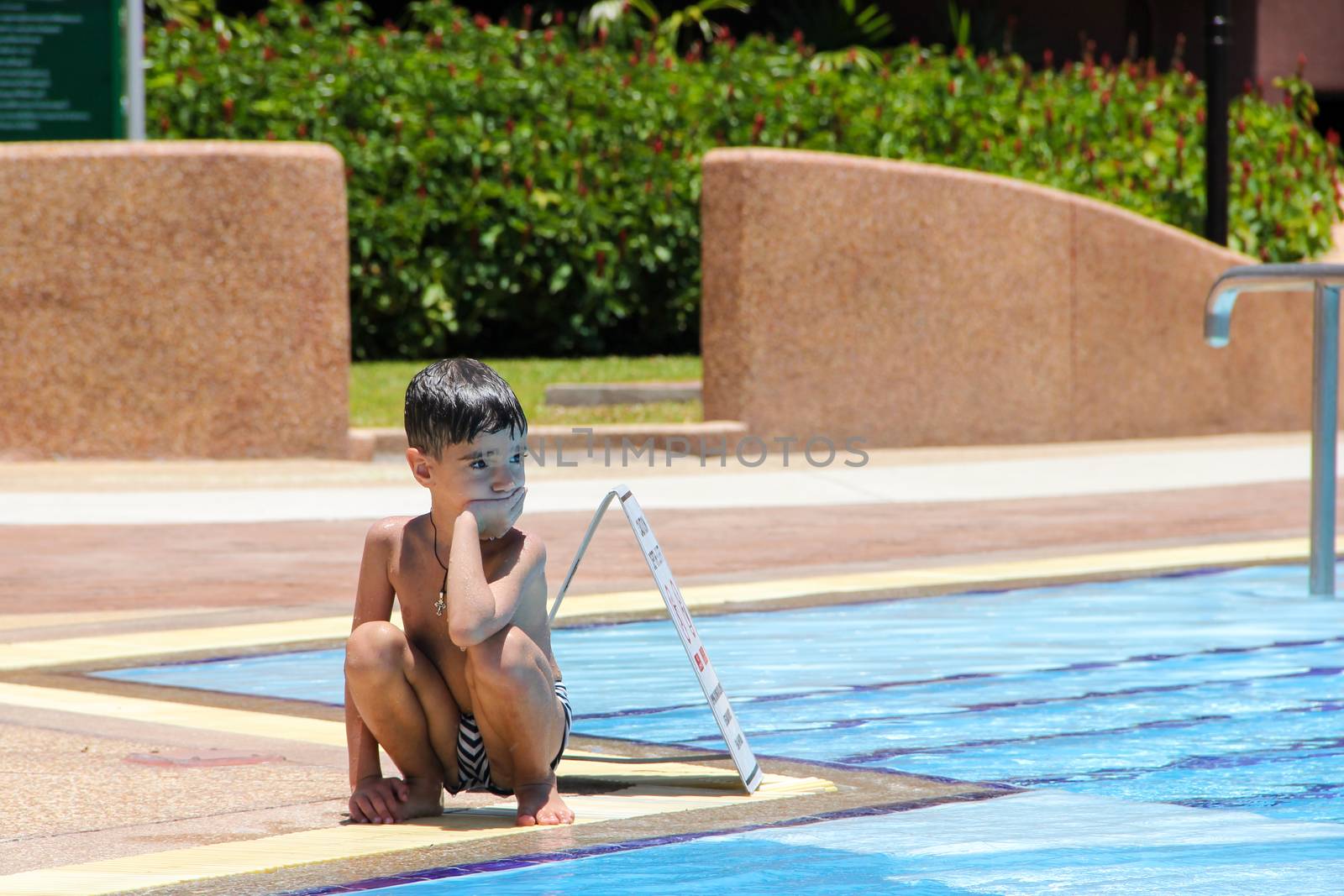 The little frustrated boy sits in swimming trunks near the swimming pool on a sunny summer day.