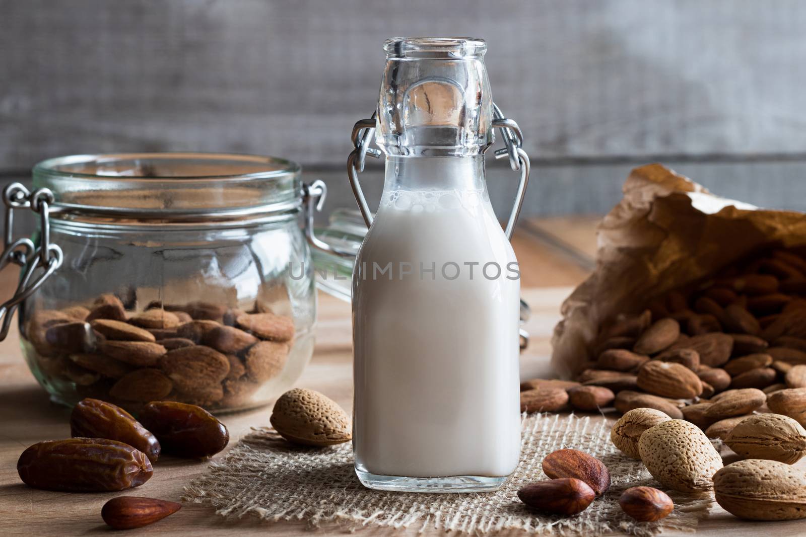 A bottle of almond milk on a wooden table, with whole (unshelled), shelled and soaked almonds and dates in the background