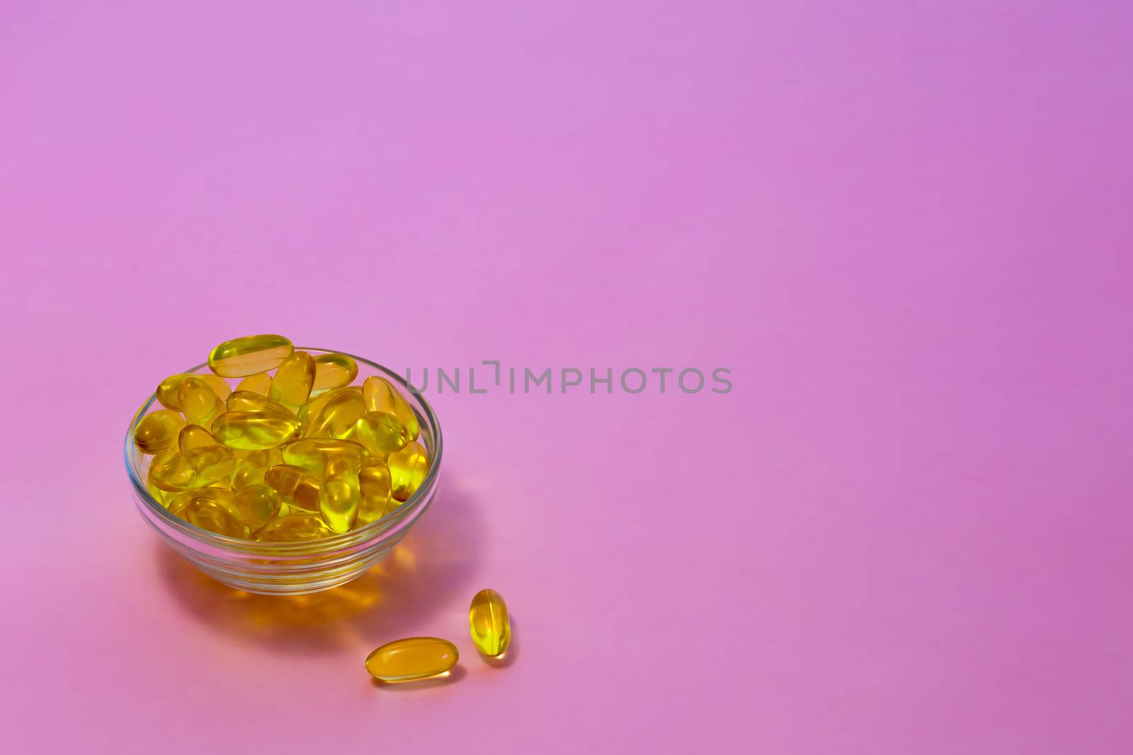 Yellow transparent capsules with fish oil containing Omega-3 acids in a transparent cup on a pink background.