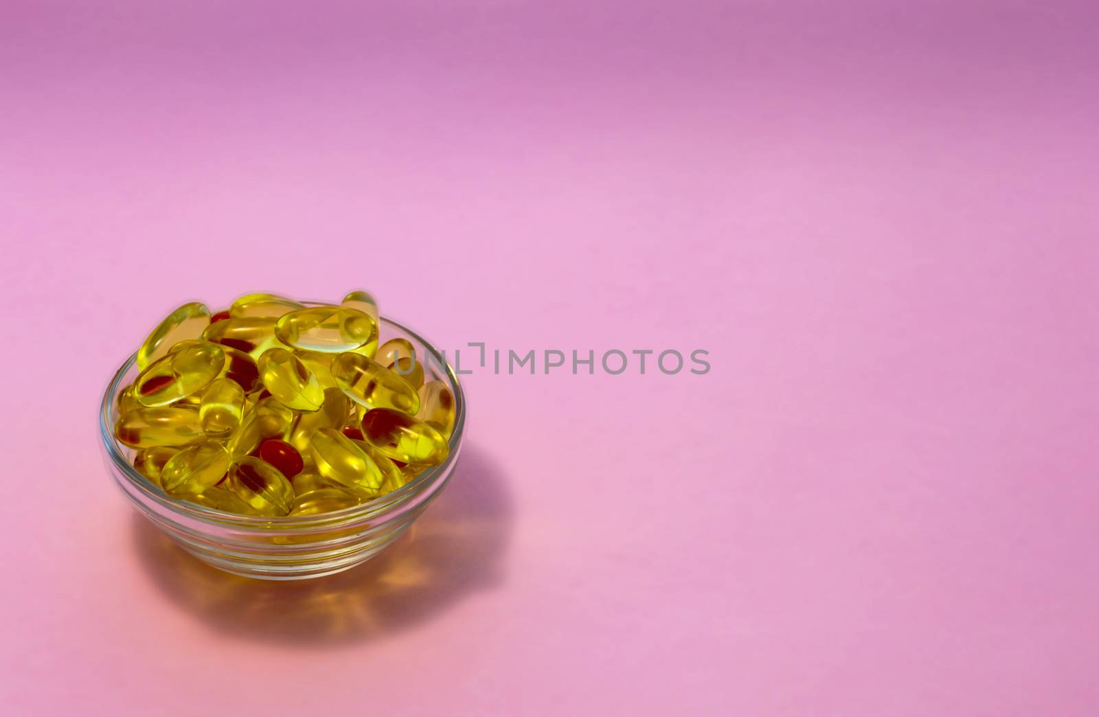 Yellow transparent capsules with fish oil containing Omega-3 acids in a transparent cup on a pink background.