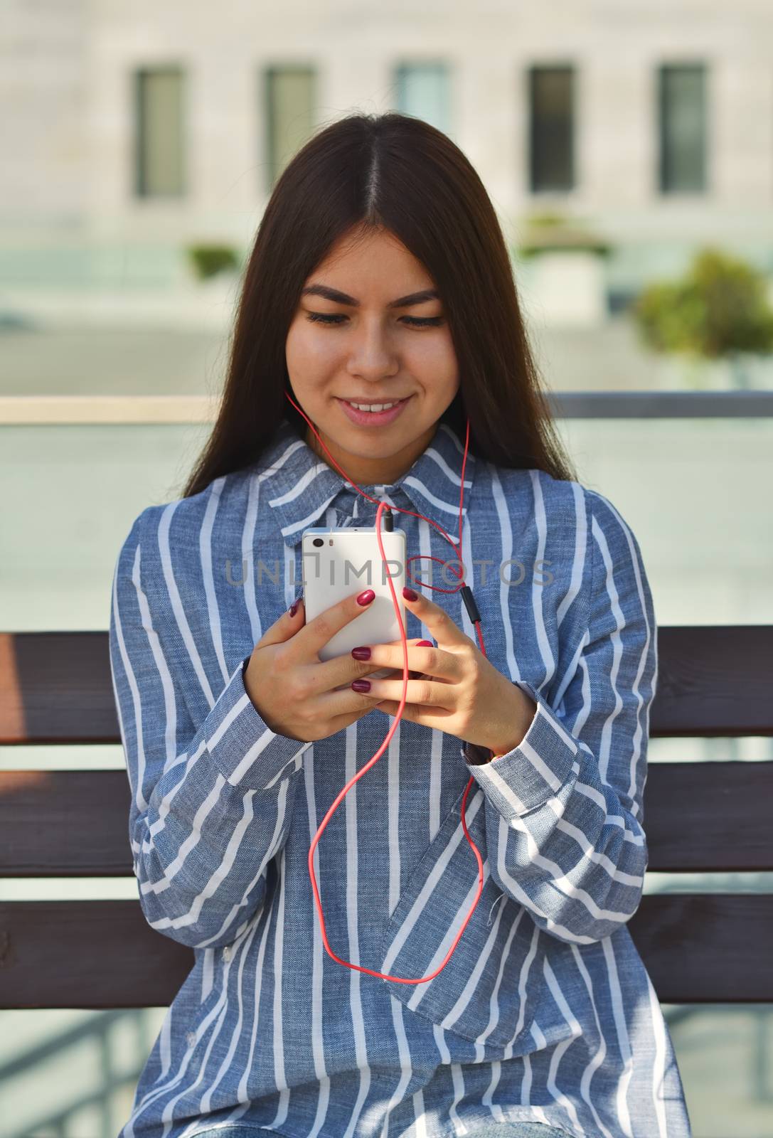 Portrait of a young beautiful brunette girl who listens to music on the street sitting on a bench