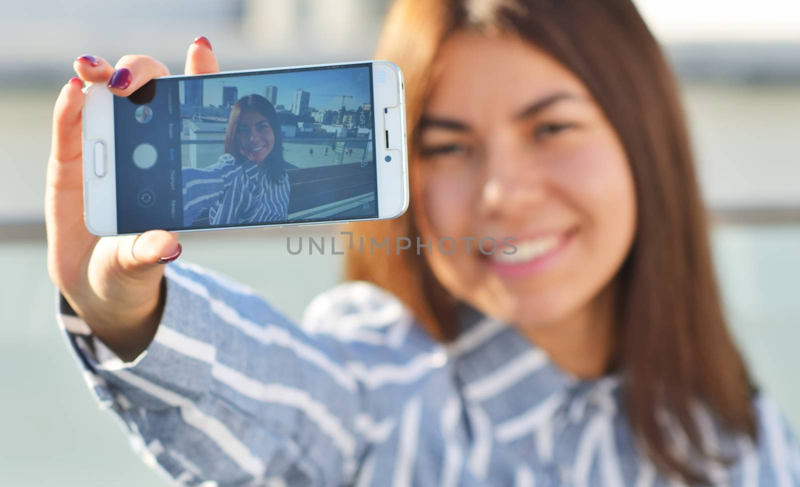 A beautiful girl in a striped blue shirt takes a selfie in the city while walking