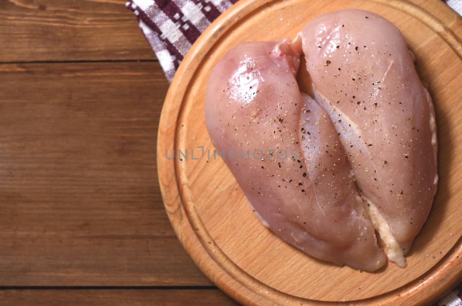 Raw chicken breast sprinkled with black pepper and salt lies on a round wooden Board. Horizontal close-up photo