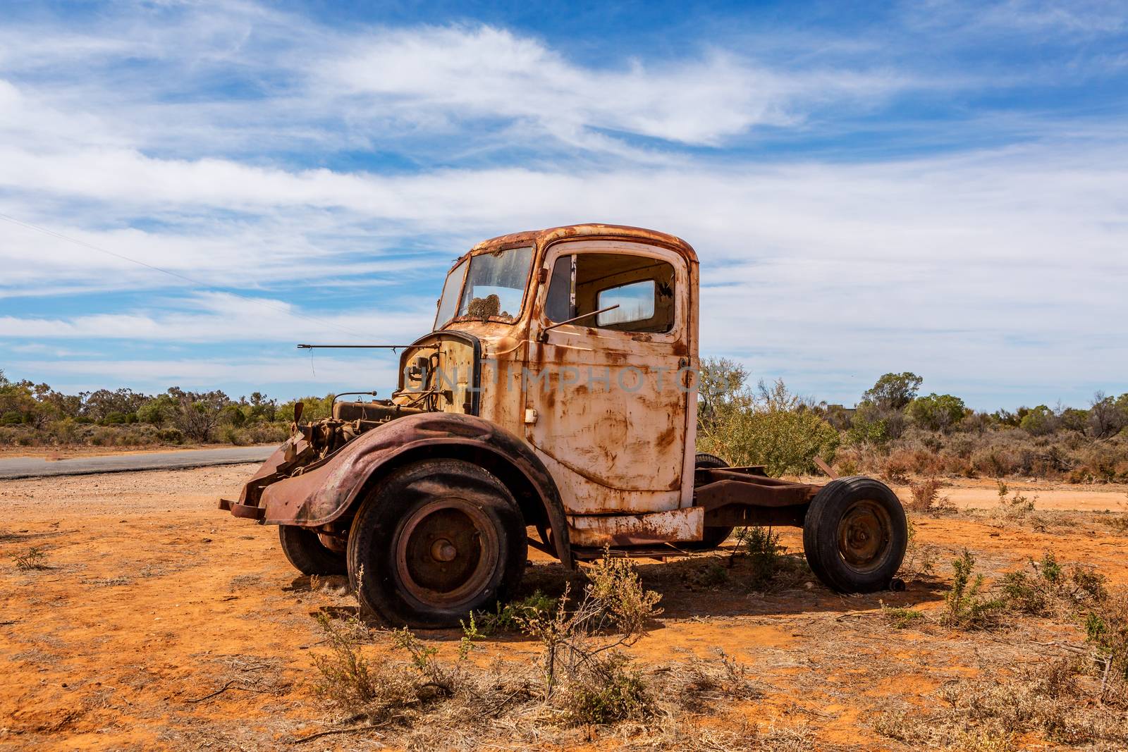 Old relic truck  metal fan sticking out from its bonnet, rusting metal panels, sits at the side of the road on red desert soils under blue skies