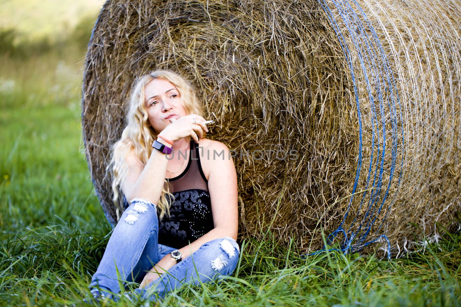 Blonde girl with long curly hair sits leaning against a haystack and smokes a cigarette