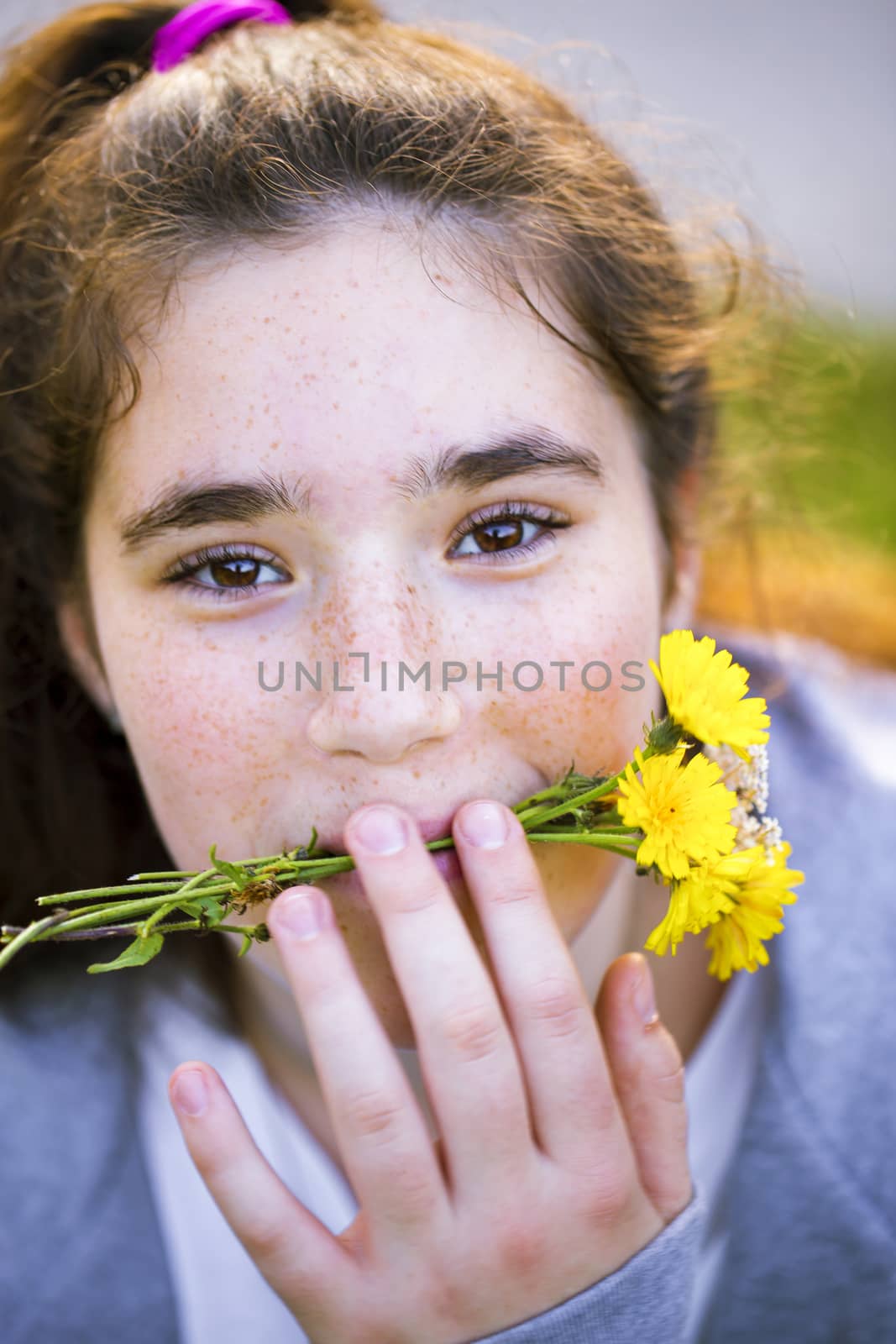 Close up portrait of a cheerful teen girl with long brown hair fooling around and holding yellow dandelions in her mouth. She is cute and funny.