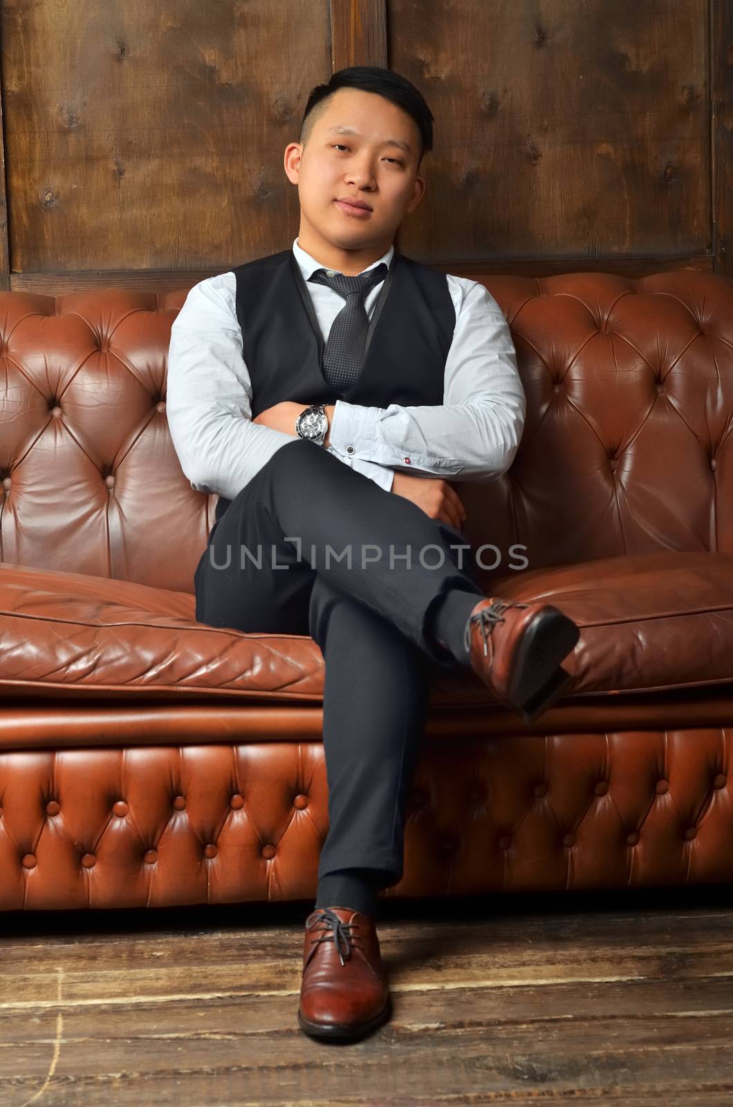 A successful Asian man sits on a leather couch, putting his foot on his leg and crossing his hands