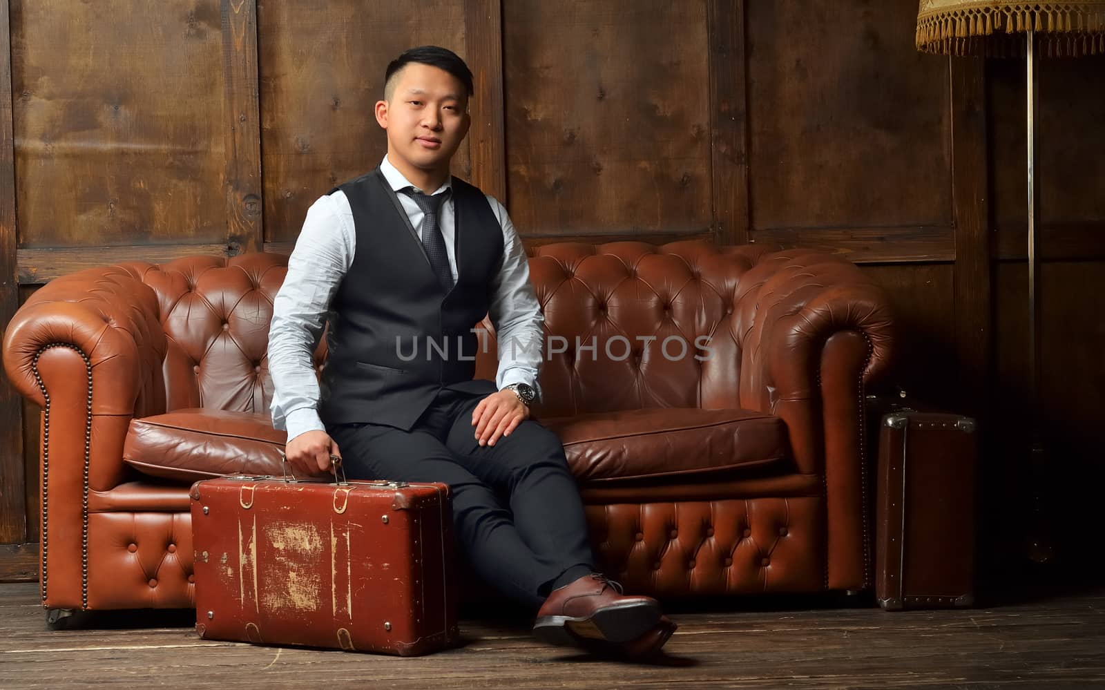 A young man in a suit sits on a leather brown sofa with a suitcase