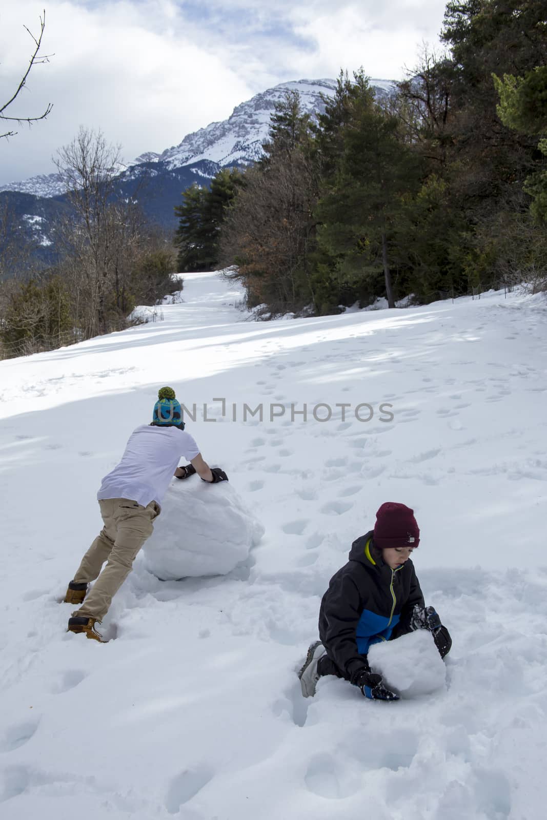 A young guy in a T-shirt and a cap makes a snowman with his younger brother in the winter forest on a nice sunny day.