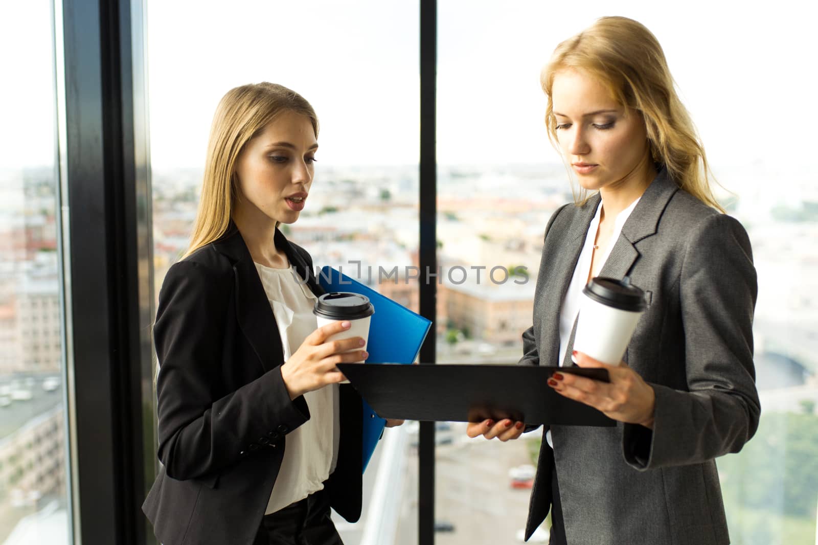 Business women discuss documents and drink coffe in office in front of panoramic windows with view at city