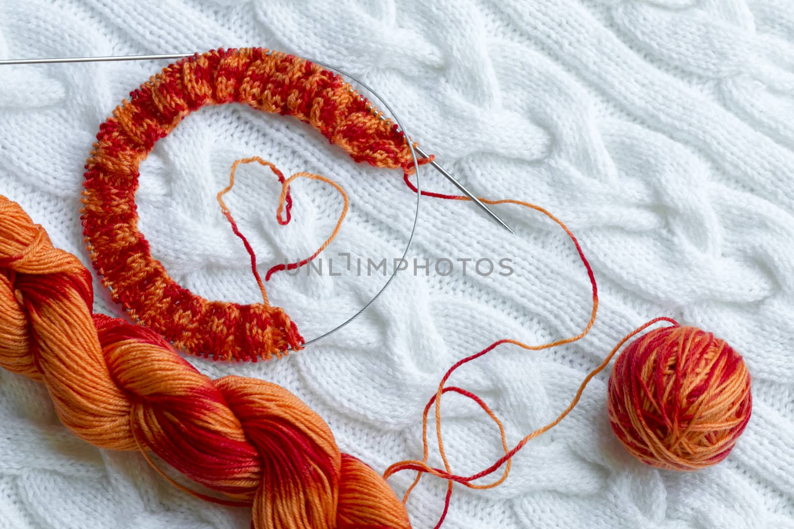 Knitting project in progress. A piece of knitting with a ball and a skein of red-orange color of sectional dyeing and knitting on the background of a white knitted plaid.