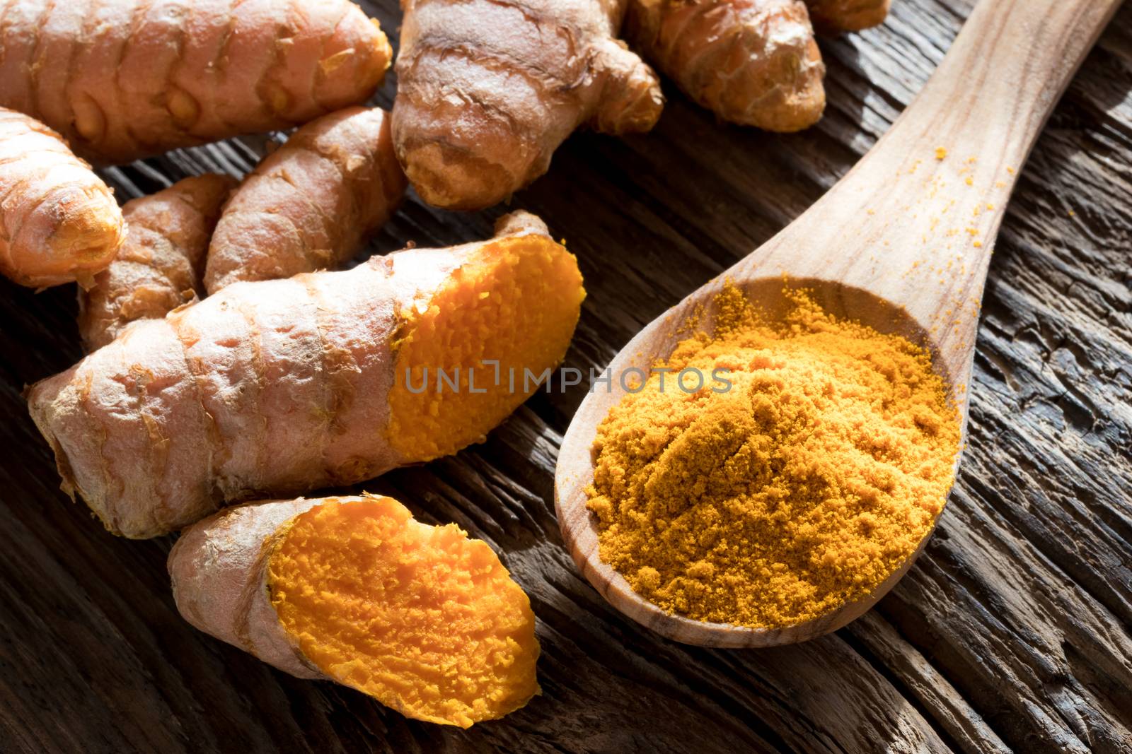 Ground turmeric on a wooden spoon, with fresh turmeric root in the background