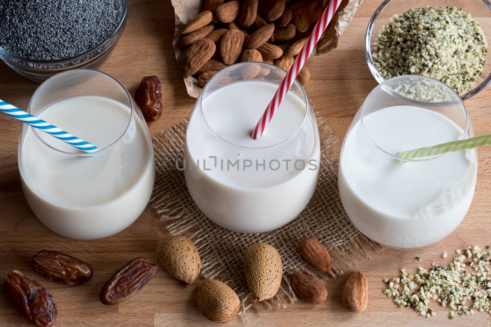 Three glasses of vegan plant milk - almond milk, poppy seed milk and hemp seed milk, with shelled and unshelled almonds, poppy seeds, hemp seeds and dates in the background