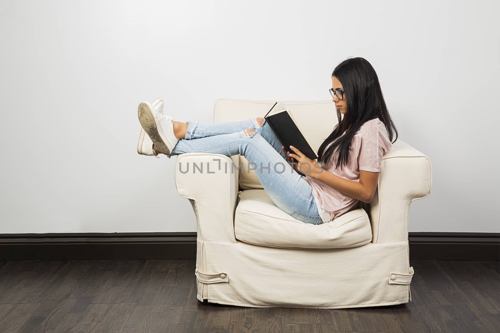 Twenty something woman sitting on the arm of a couch, with her feet up, reading a book