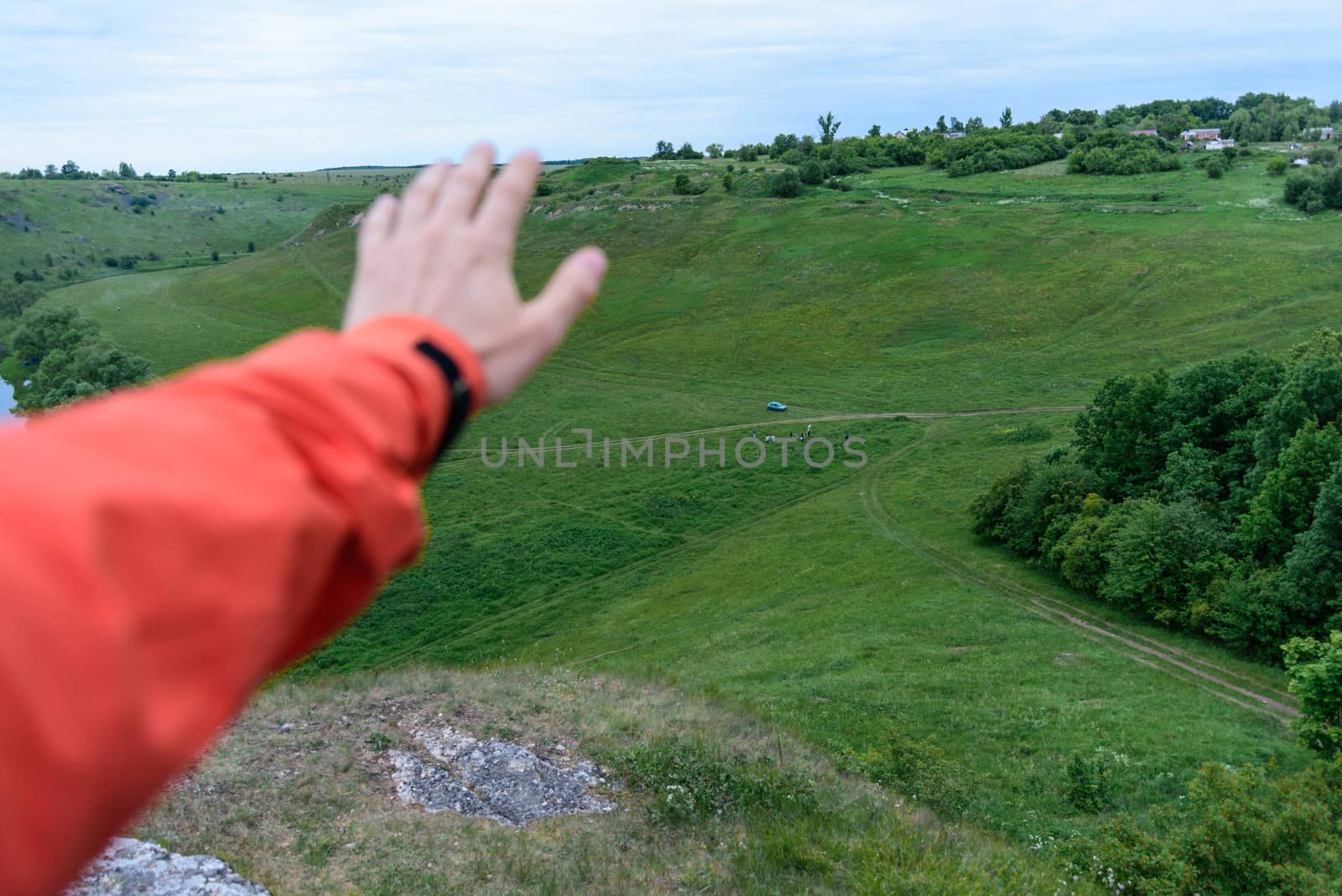 Man traveler in an orange jacket inspects the neighborhood, showing his hand on the landscape, selective focus.