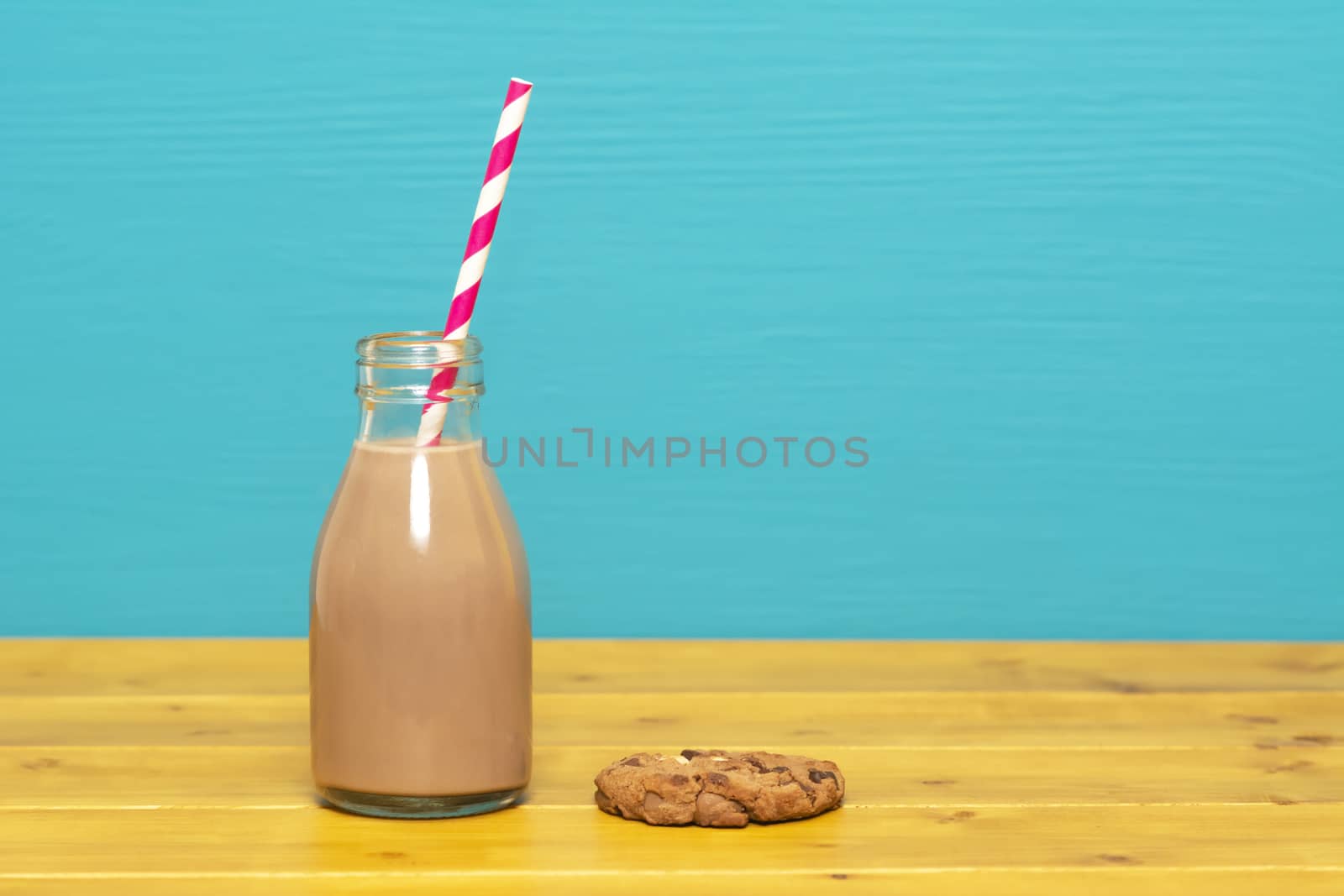 Chocolate milkshake with a retro paper straw in a one-third pint glass milk bottle and a chocolate chip cookie, on a wooden table against a teal background