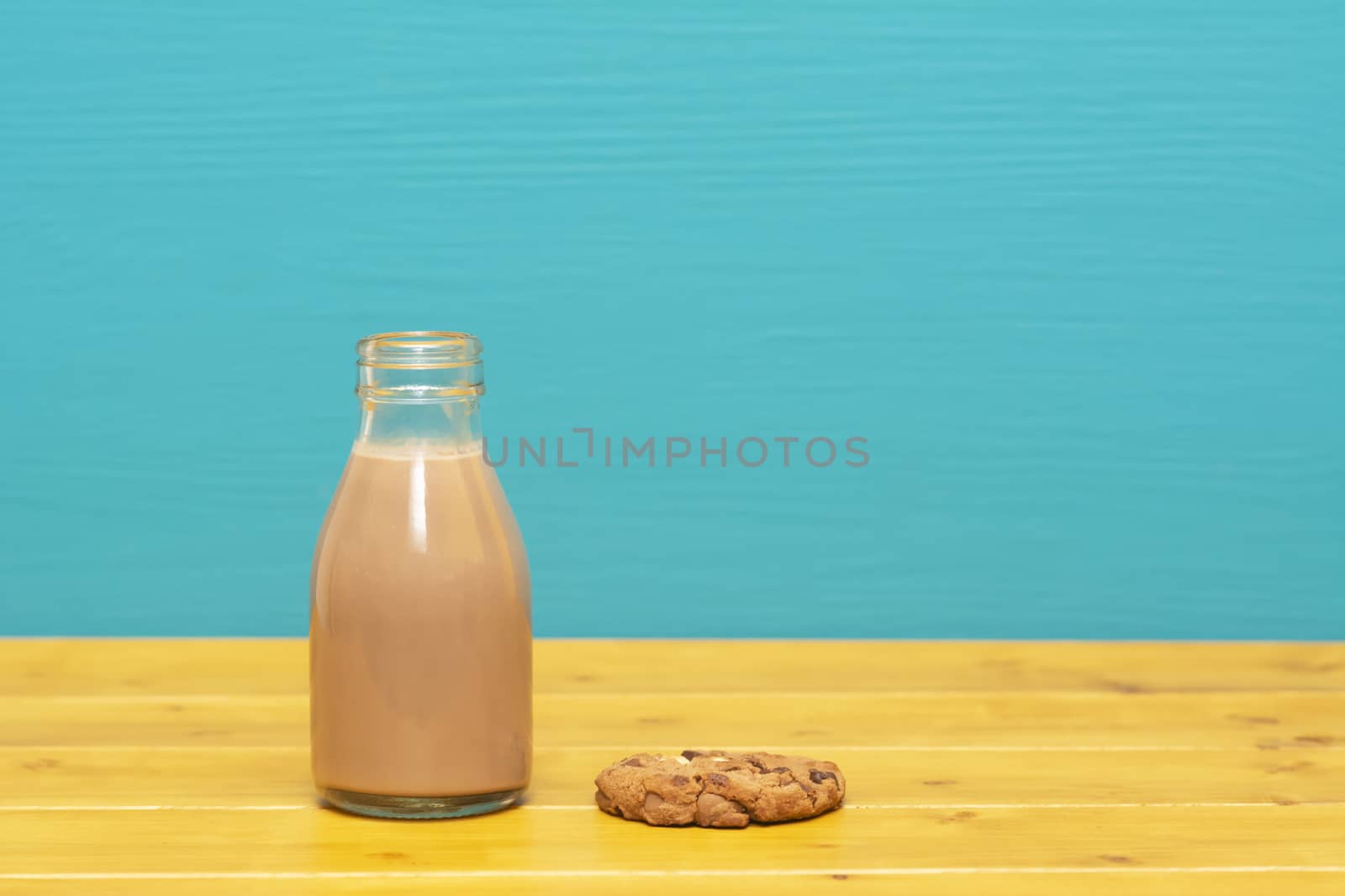 Chocolate milkshake in a retro one-third pint glass milk bottle and a chocolate chip cookie, on a wooden table against a bright teal painted background