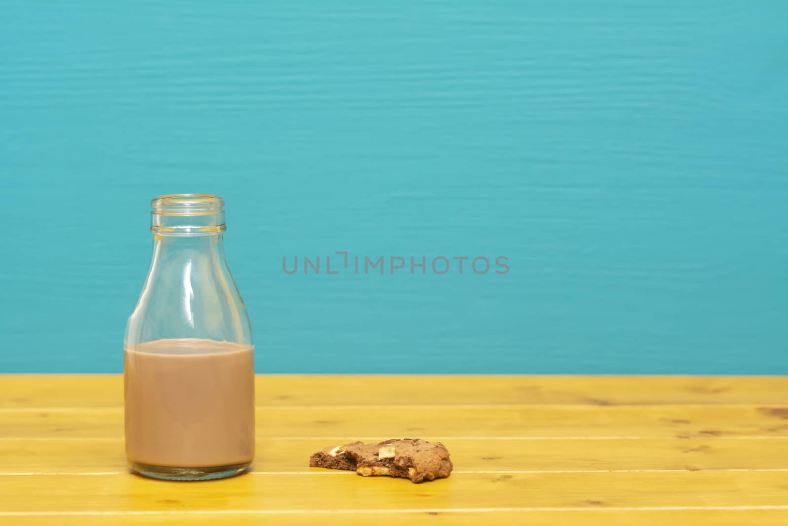 One-third pint glass milk bottle half full with chocolate milkshake and a half-eaten chocolate chip cookie, on a wooden table against a teal background