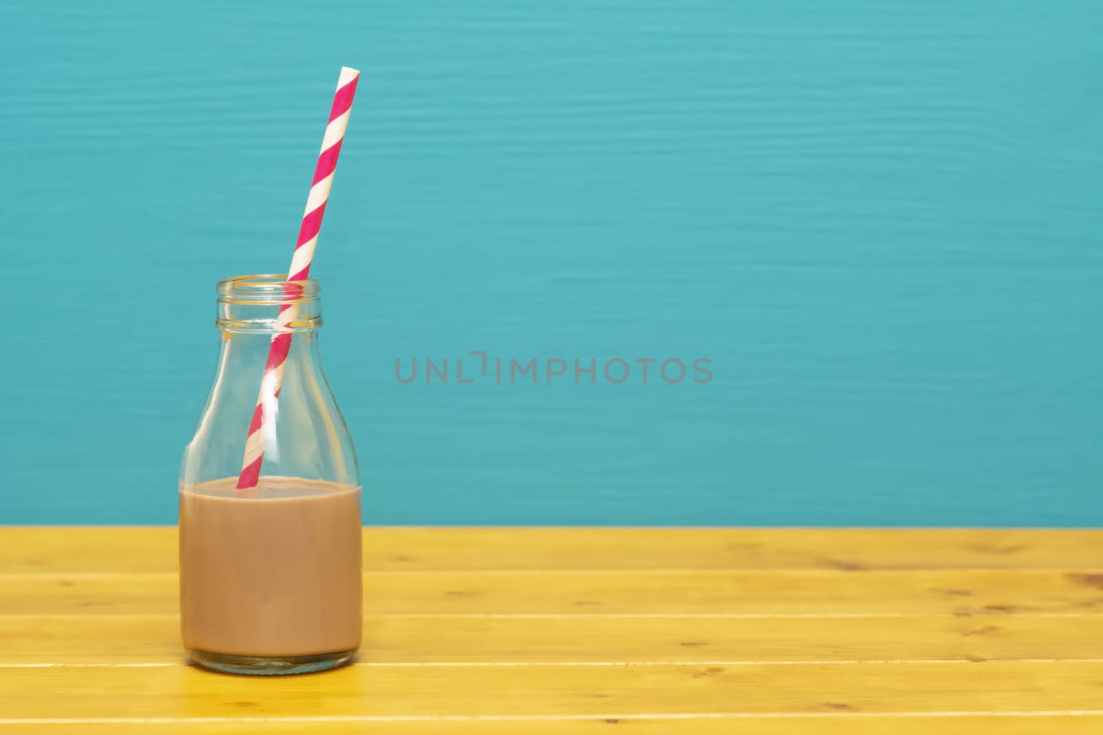 One-third pint glass milk bottle half full with chocolate milkshake with a retro paper straw, on a wooden table against a teal background