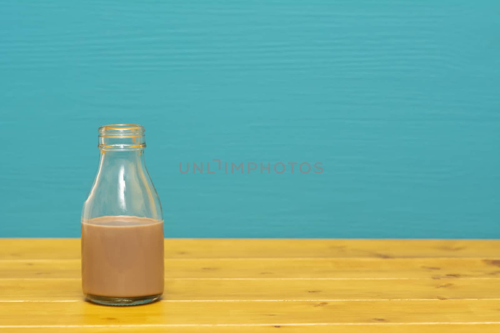 One-third pint glass milk bottle half full with chocolate milkshake, on a wooden table against a bright teal painted background