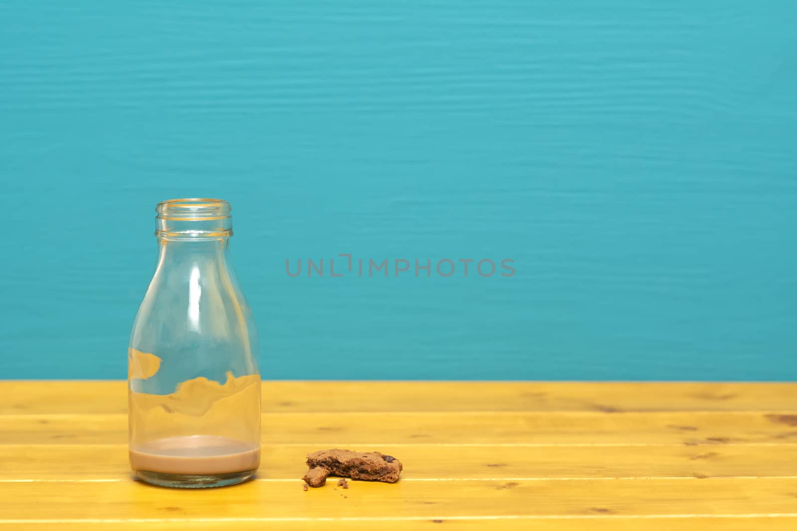 One-third pint glass milk bottle with dregs of chocolate milkshake and chocolate chip cookie crumbs, on a wooden table against a teal background