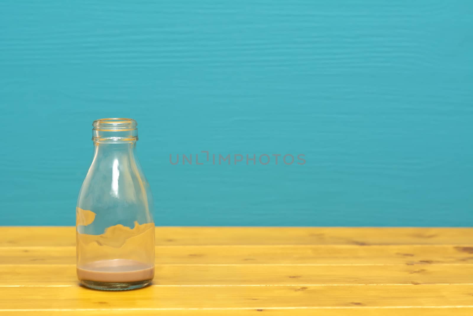 One-third pint glass milk bottle with dregs of chocolate milkshake, on a wooden table against a bright teal painted background
