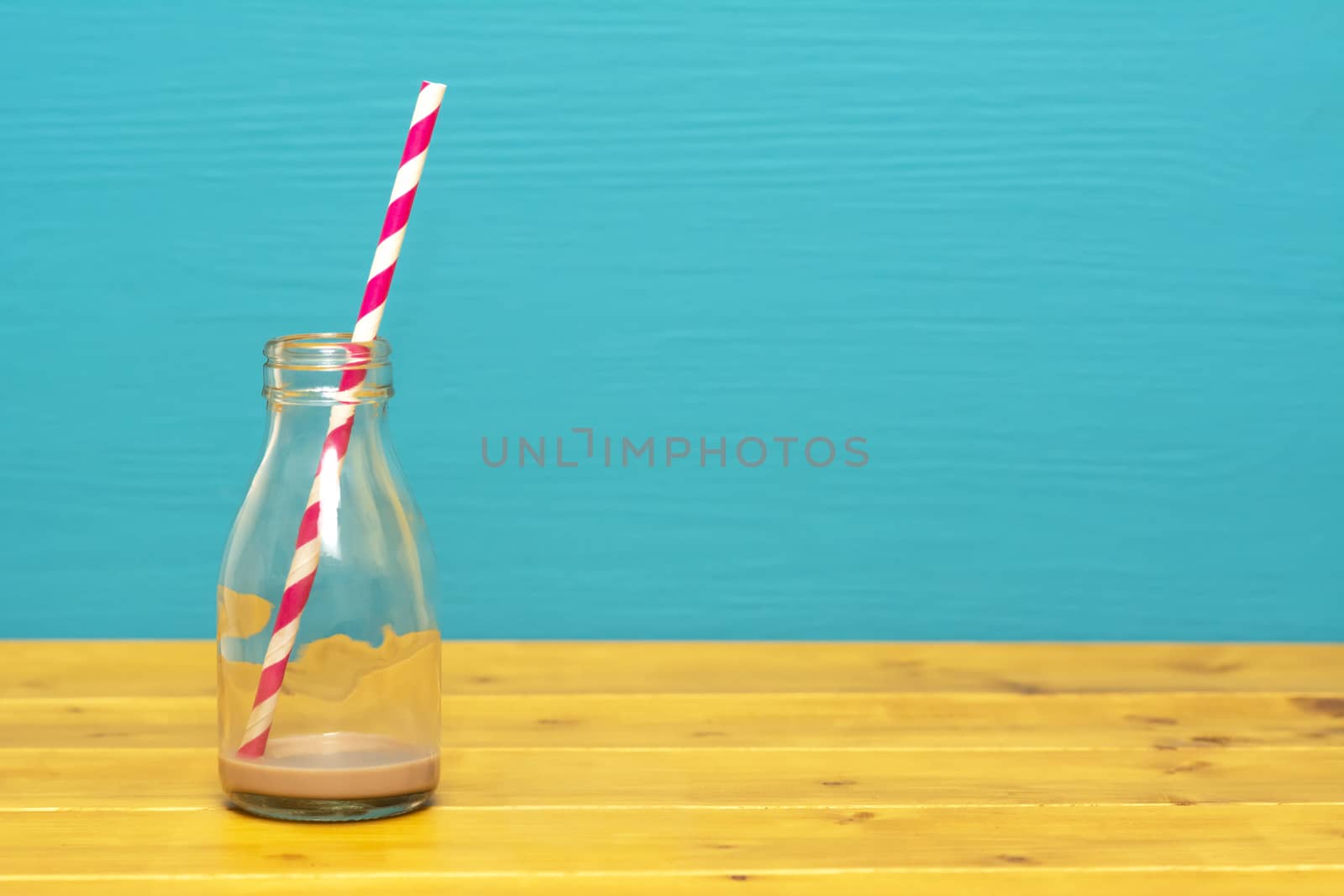 One-third pint glass milk bottle with dregs of chocolate milkshake with a retro paper straw, on a wooden table against a teal background