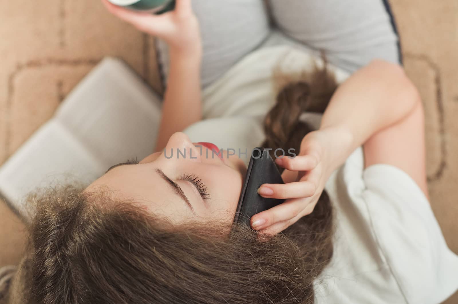 Cheerful brunette girl talking on mobile phone while sitting on comfortable couch. Smiling hipster girl having cell telephone conversation while resting on sofa in home interior and drinks coffee