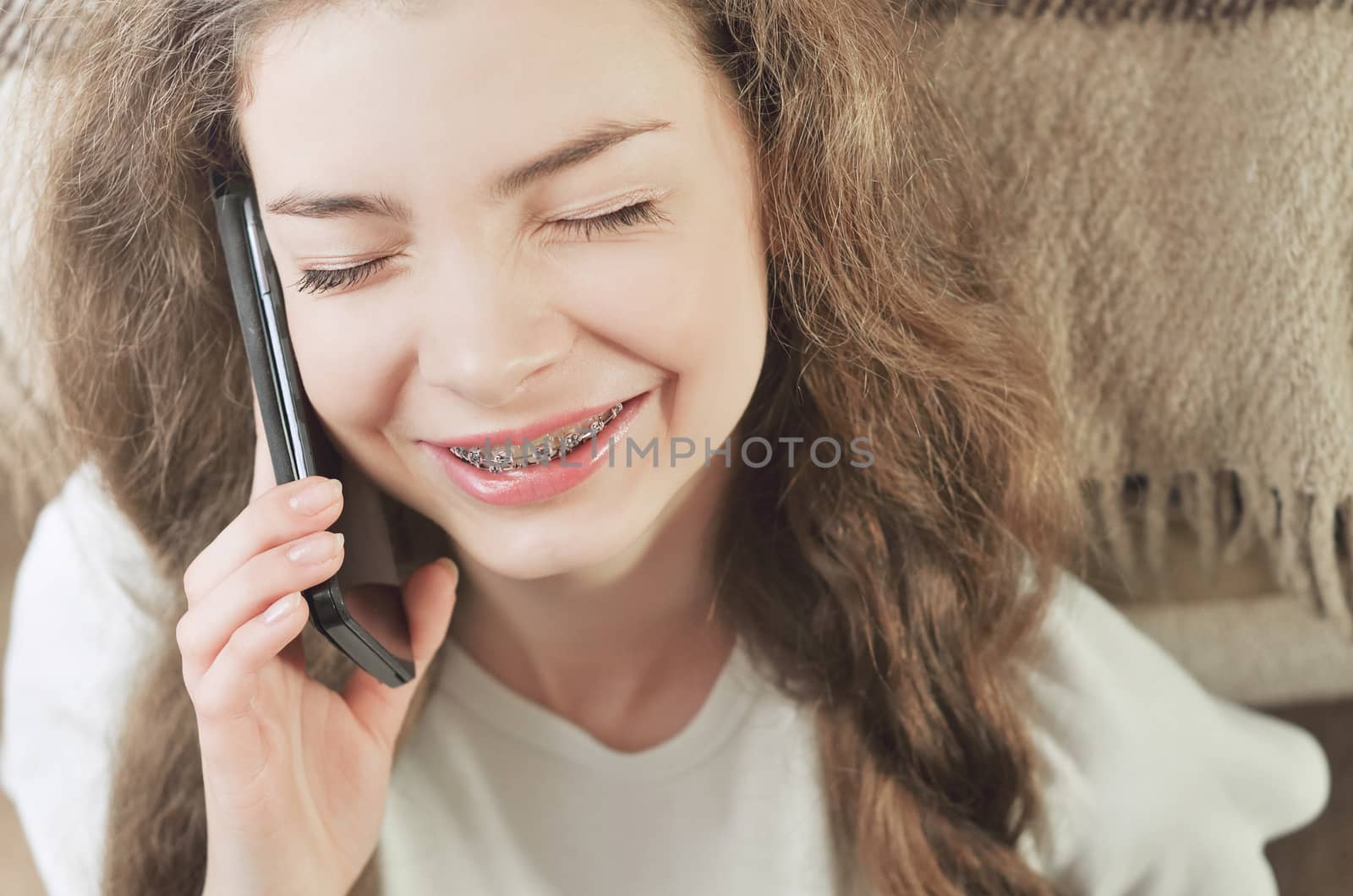 Cheerful brunette girl talking on mobile phone while sitting on comfortable couch. Smiling hipster girl having cell telephone conversation while resting near sofa