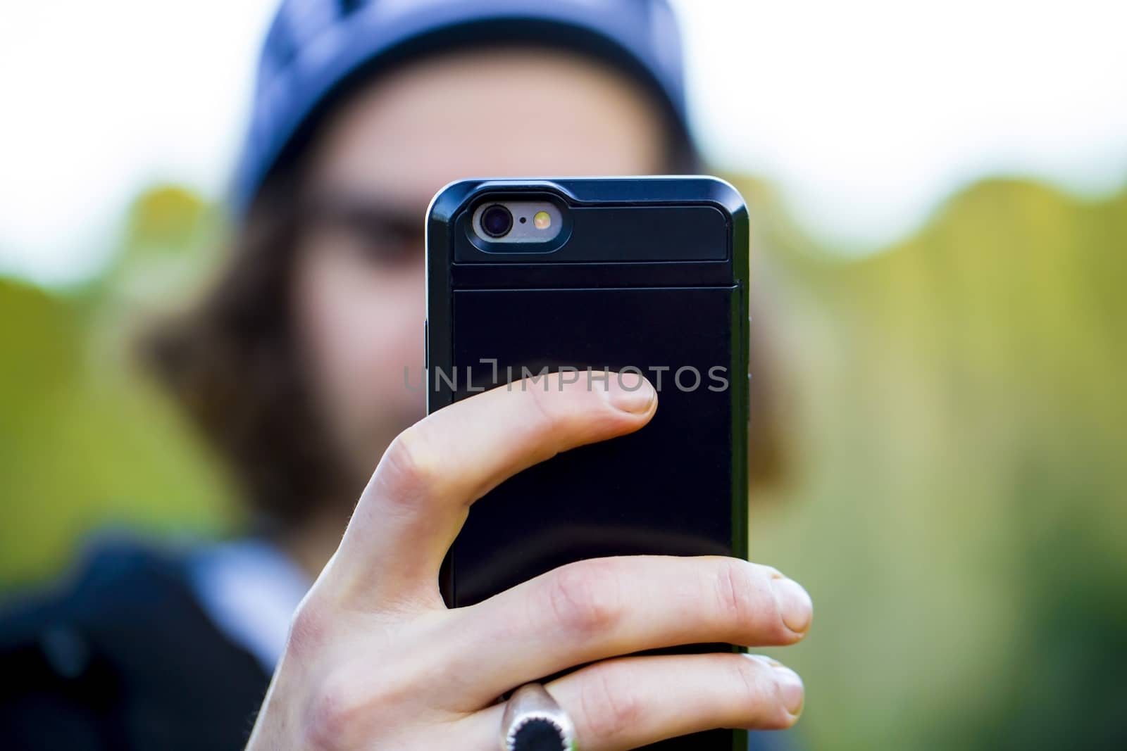 Unrecognizable man with long hair, wearing warm clothes and a cap, taking a selfie photo with his smartphone on the street in autumn. Close up on the hand that holds the mobile phone in a black case.