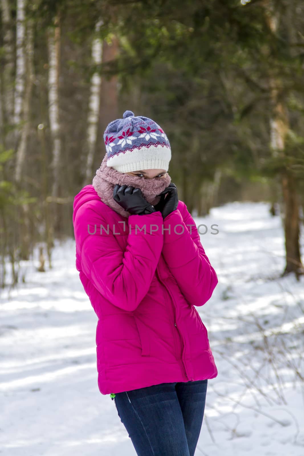 Portrait of a teenage girl in a bright pink winter jacket and knitted hat, which covers her face with a scarf. She stay in the winter snowy forest.