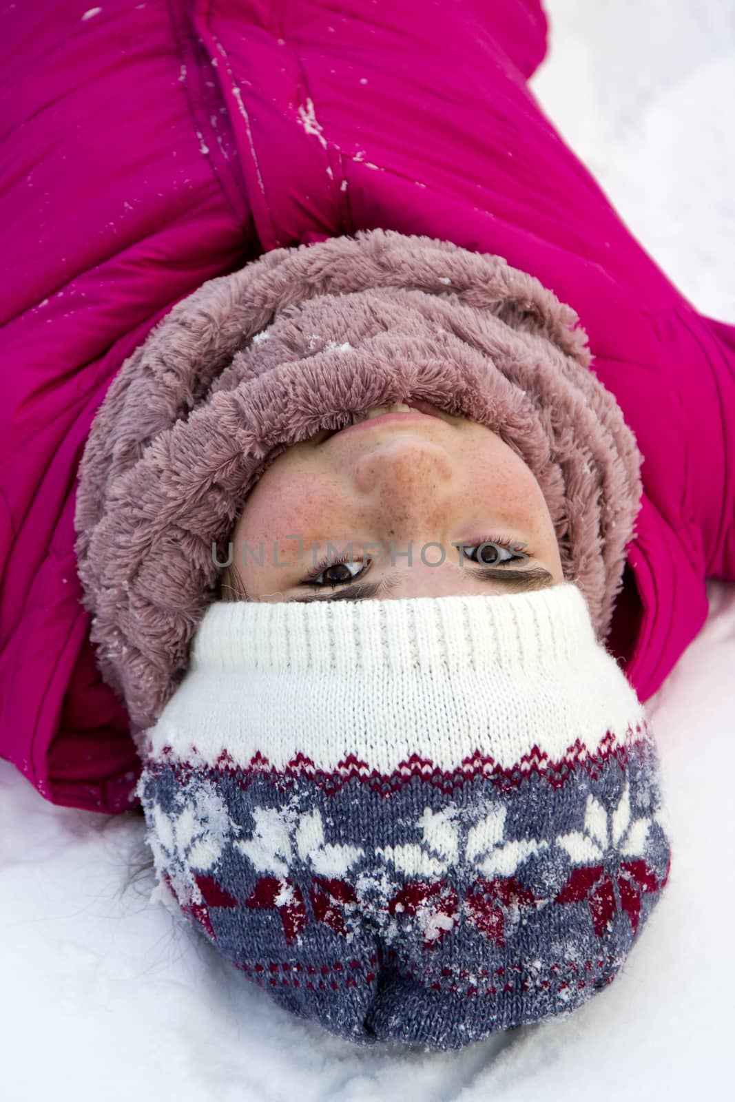 Portrait of a teenage girl in a bright pink winter jacket and knitted hat in the winter in the open air, she lies on pure white snow.