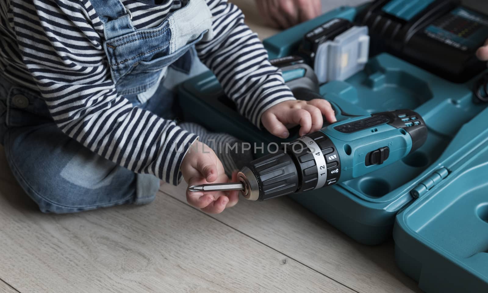 a one  year old child sitting on the ground putting a new drill in a drilling machine  which is located in a blue plastic case