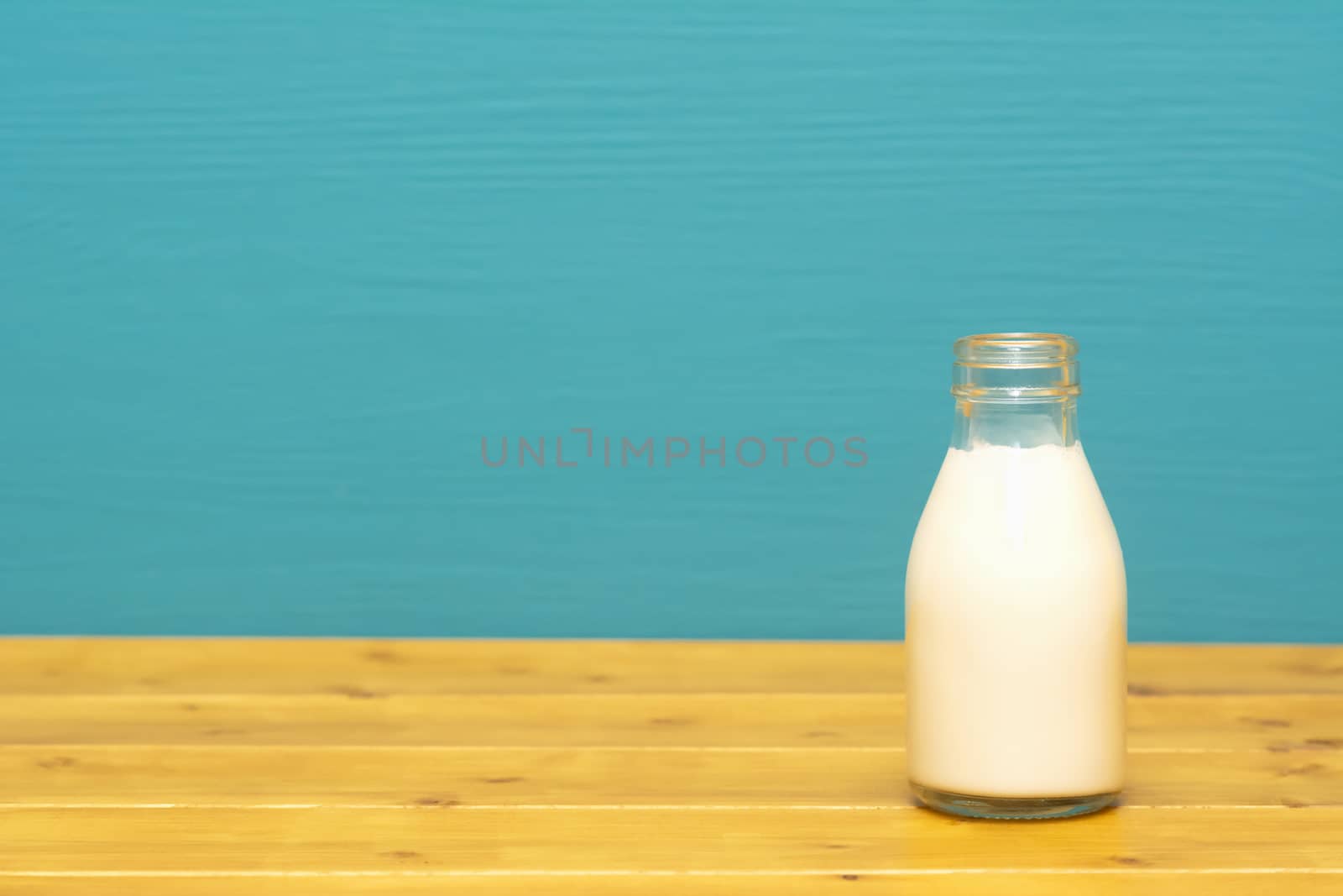 Fresh creamy milk in a one-third pint glass milk bottle, on a wooden table against a bright teal painted background