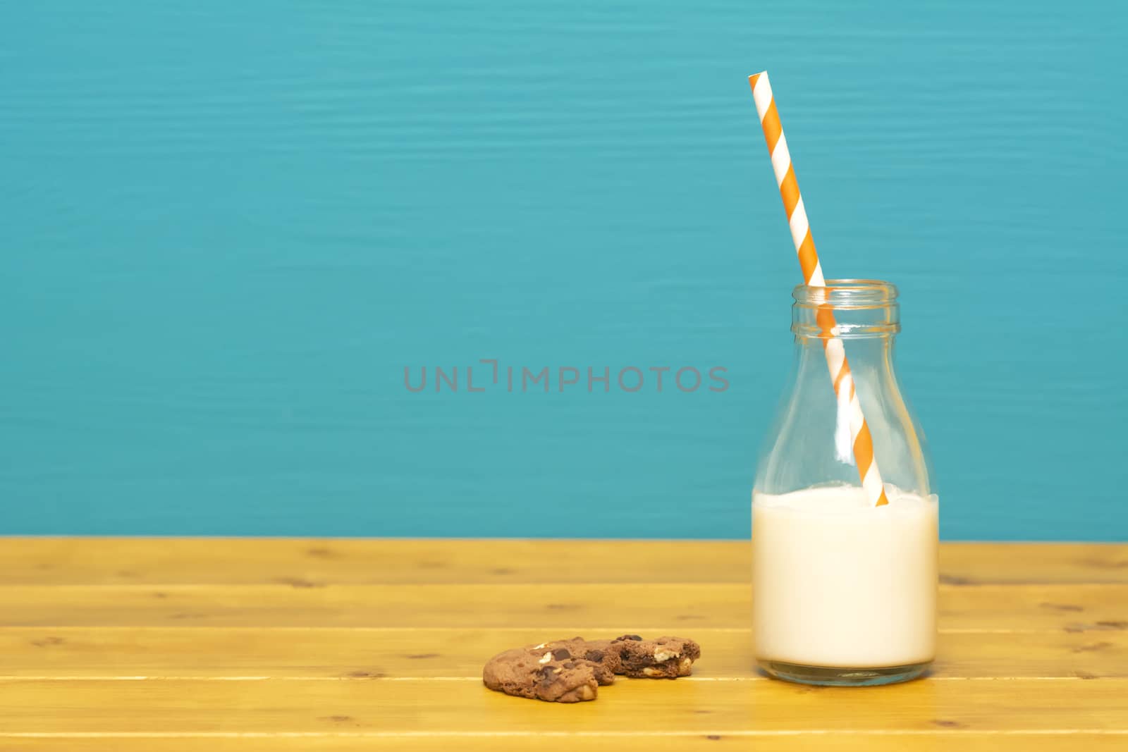 One-third pint glass milk bottle half full with fresh creamy milk with a retro straw and a half-eaten chocolate chip cookie, on a wooden table against a teal background