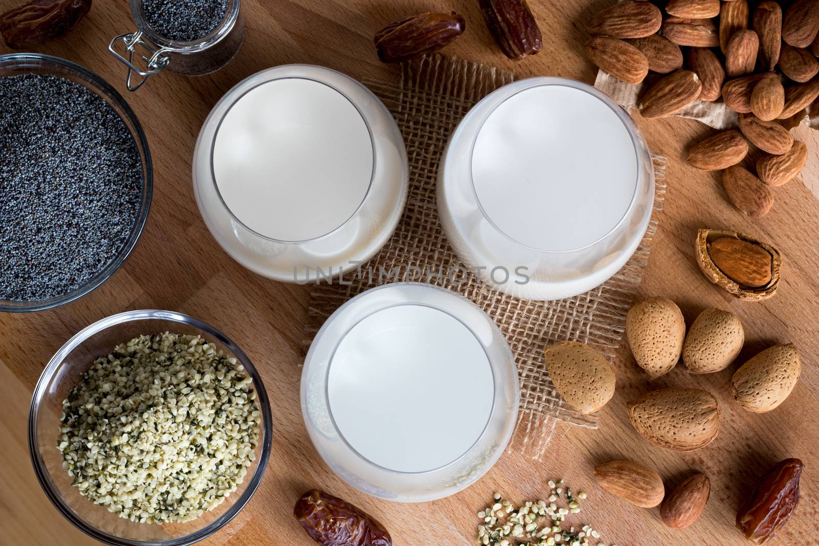 Top view of three glasses of vegan plant milk - almond milk, poppy seed milk and hemp seed milk, with shelled and unshelled almonds, poppy seeds, hemp seeds and dates on a wooden table