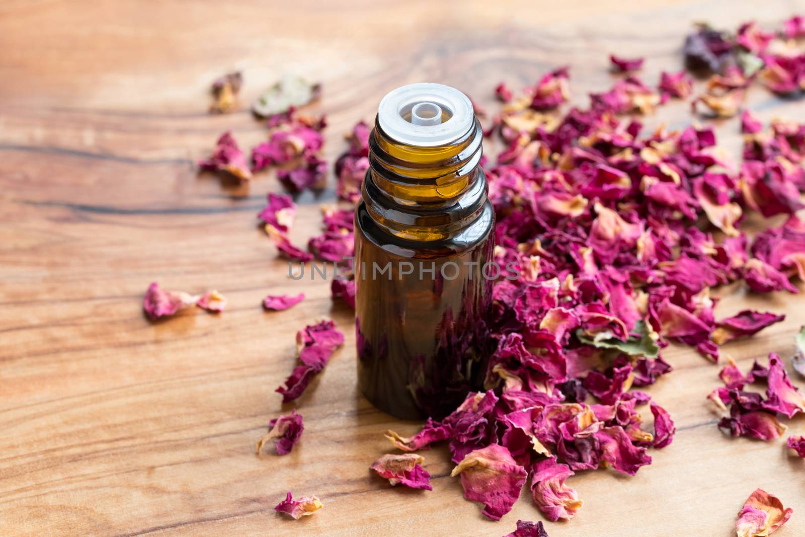 A bottle of rose essential oil with dried rose petals on a wooden table