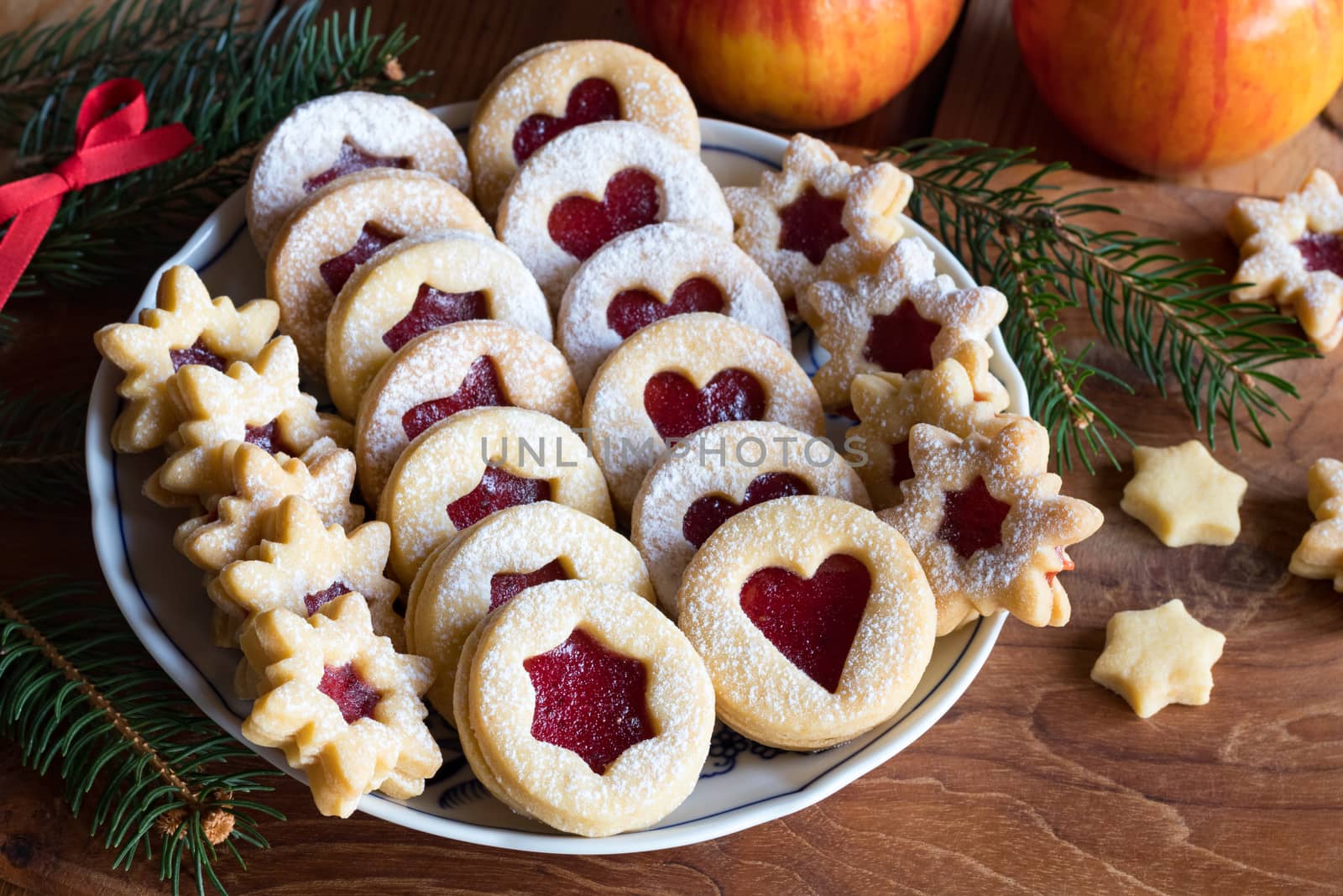 Linzer Christmas cookies filled with strawberry jam and dusted with sugar, arranged on a plate on a wooden table, with apples and spruce branches in the background