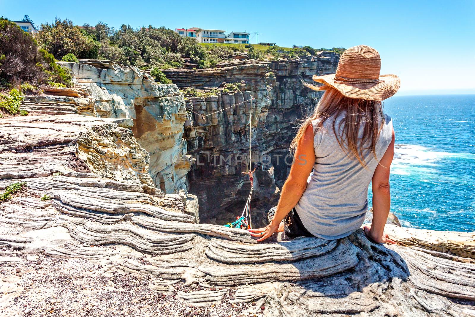 female sitting cliffside watching highliners daringly walk the rope high above the ocean