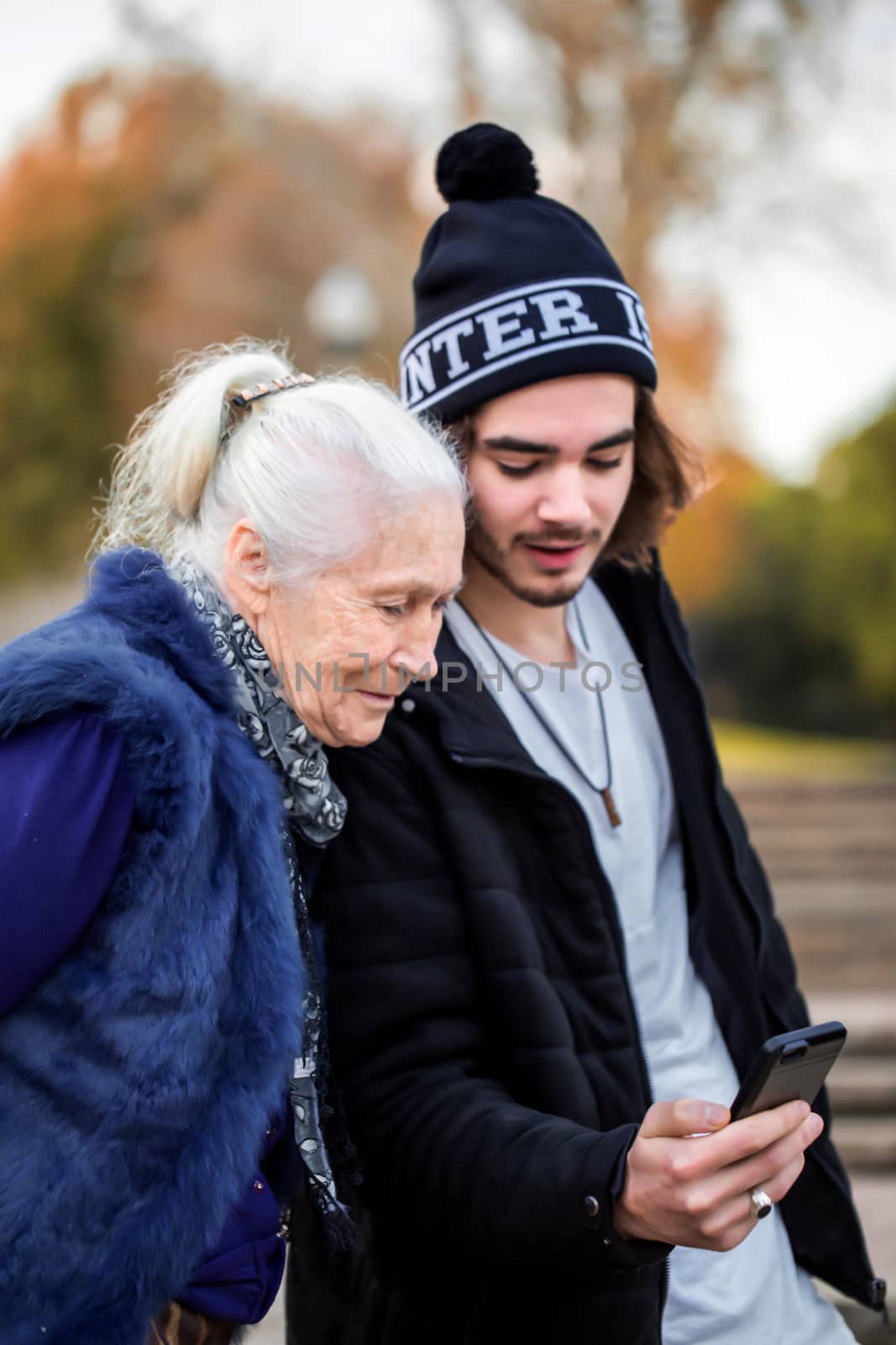 Pleasant young man explains to his elegant gray-haired grandmother how to use a smartphone. They walk in the autumn park and wear casual clothes.