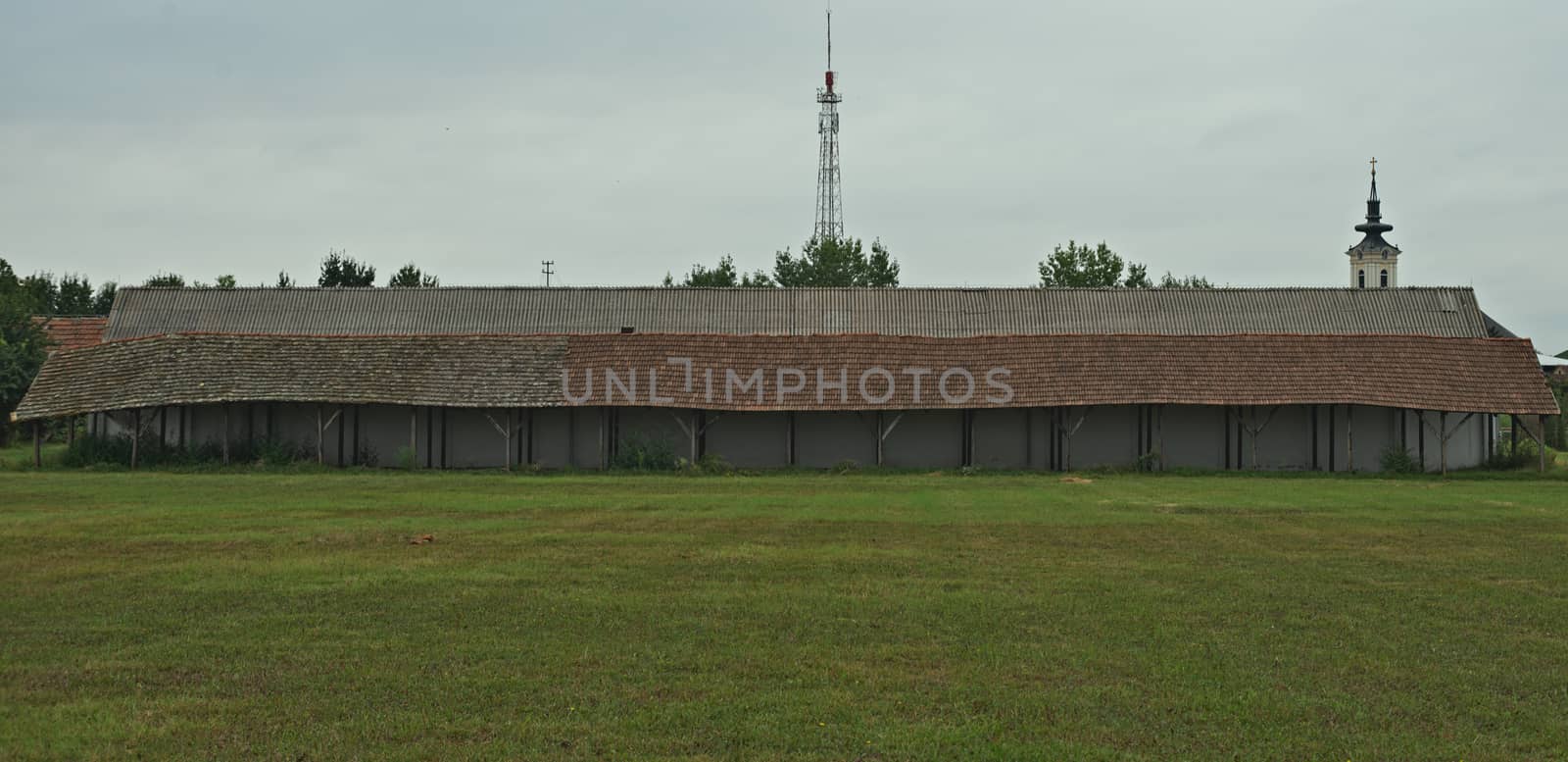 Long shed next to stable on a large field with church and radio tower in background