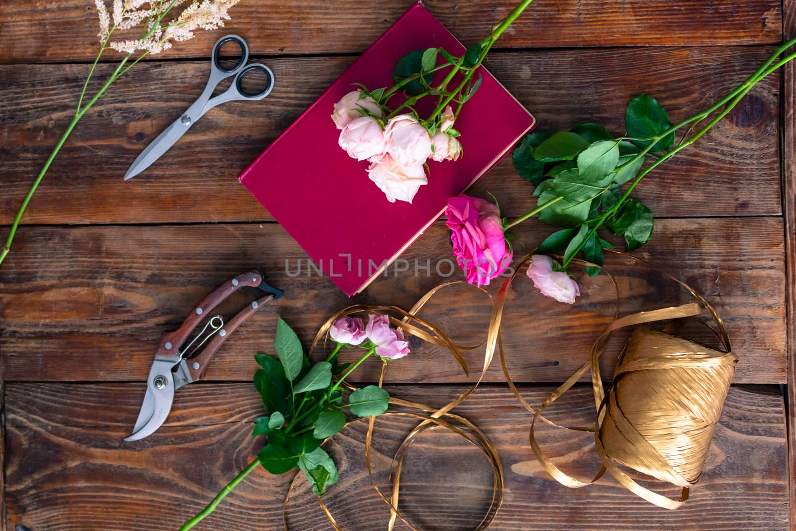 On the wooden surface laid out tools for flower bouquets: scissors, roses, colored ribbon, roses.