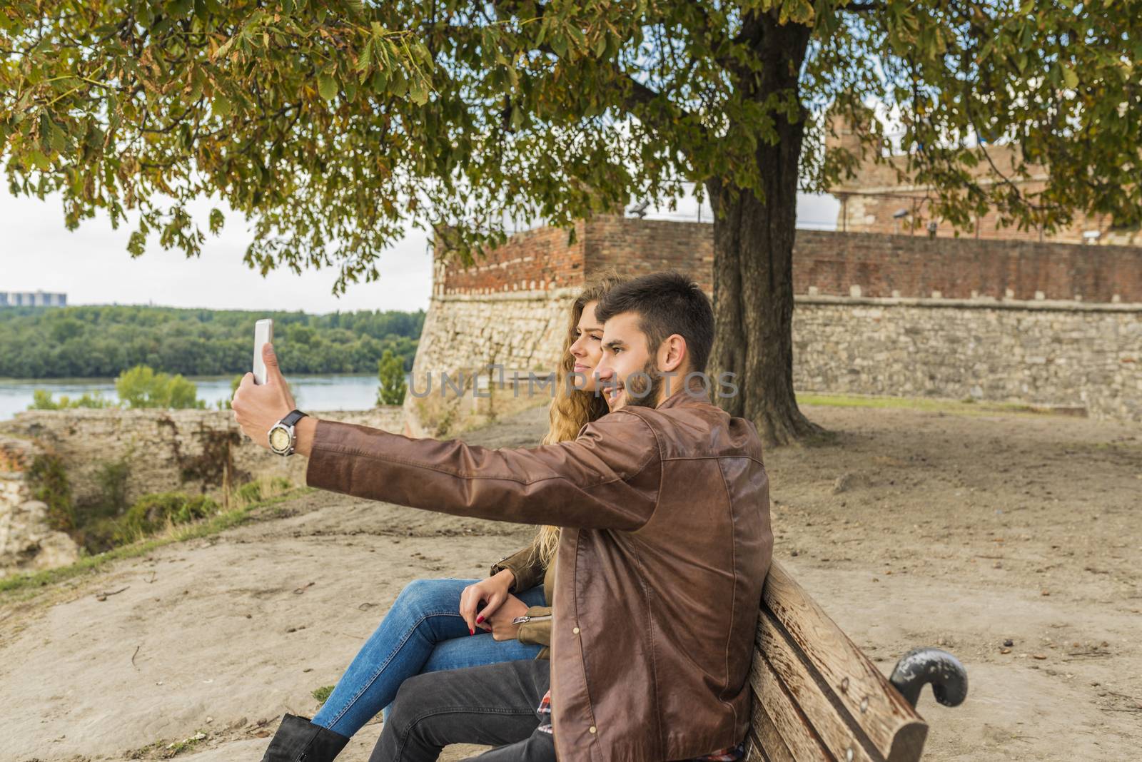 Young man is taking selfie with his beautiful girlfriend while sitting on the bench in the park under the tree