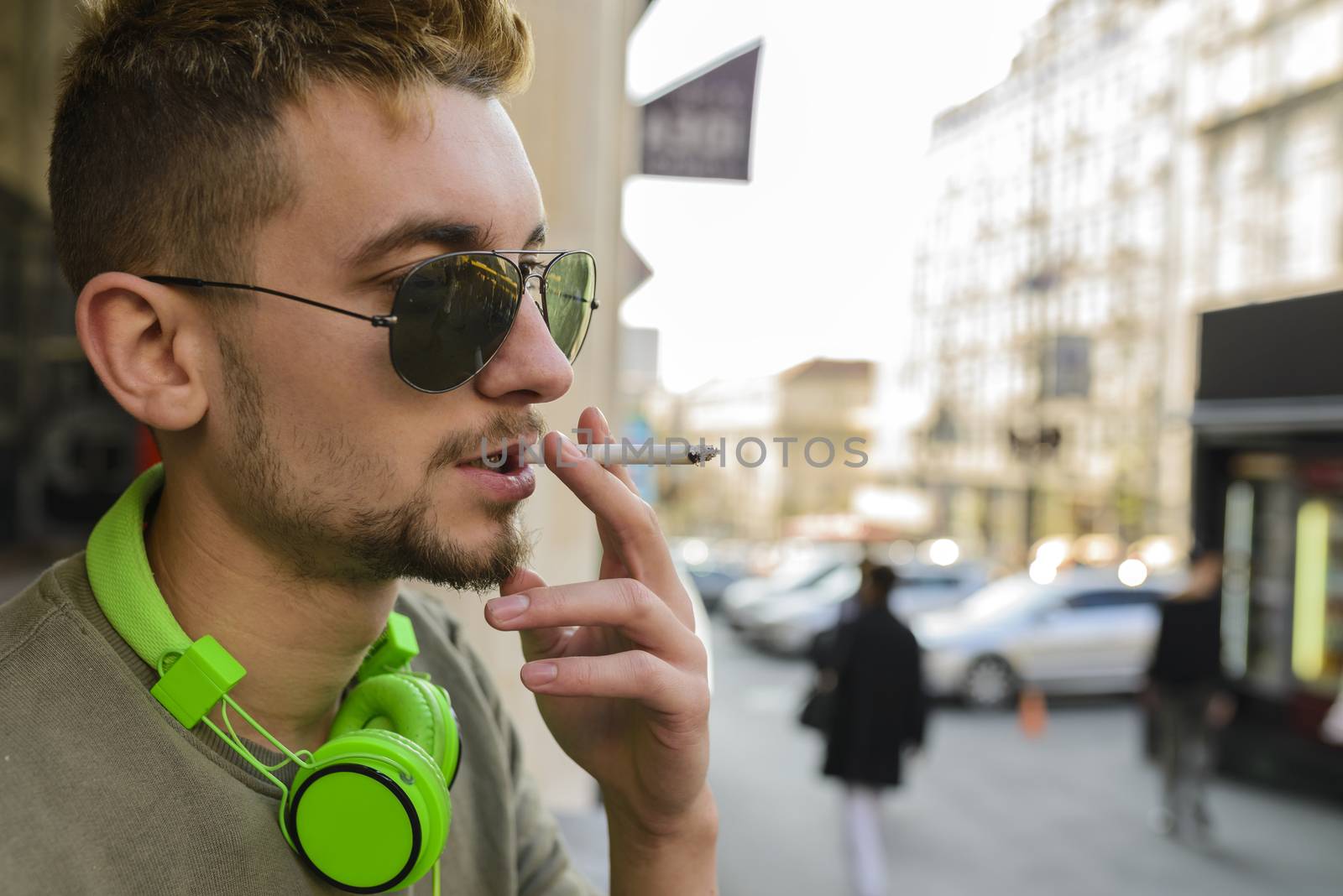 Young handsome man with green headphones and sunglasses enjoying a cigarette in the street.